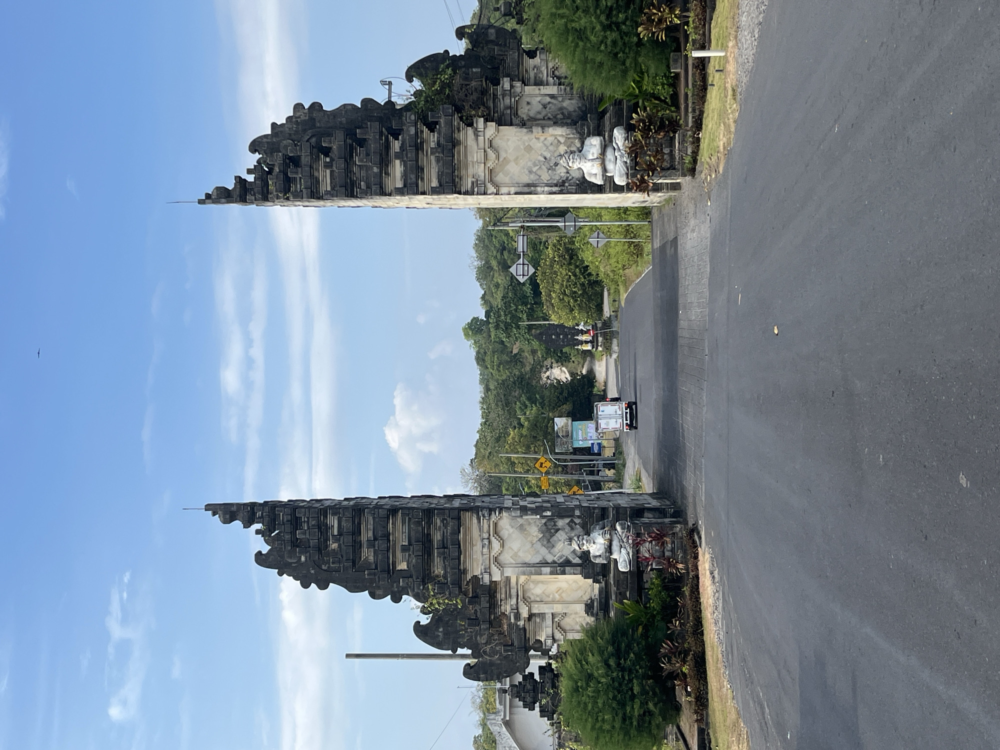 The image depicts the Candi Bentar gate, a prominent landmark in the city of Kuta Selatan, Indonesia. The gate is an ancient Balinese architectural structure, featuring two symmetrical, parallel walls adorned with intricate carvings and traditional motifs. It is situated on the side of a paved road, with lush green vegetation and trees surrounding the area. Two white statues of seated figures are placed at the base of the gate. A white truck is seen driving on the road towards the gate, disappearing into the distance. The sky above is blue with scattered white clouds, indicating a bright, sunny day. The scene conveys a sense of cultural heritage and natural beauty characteristic of Bali.