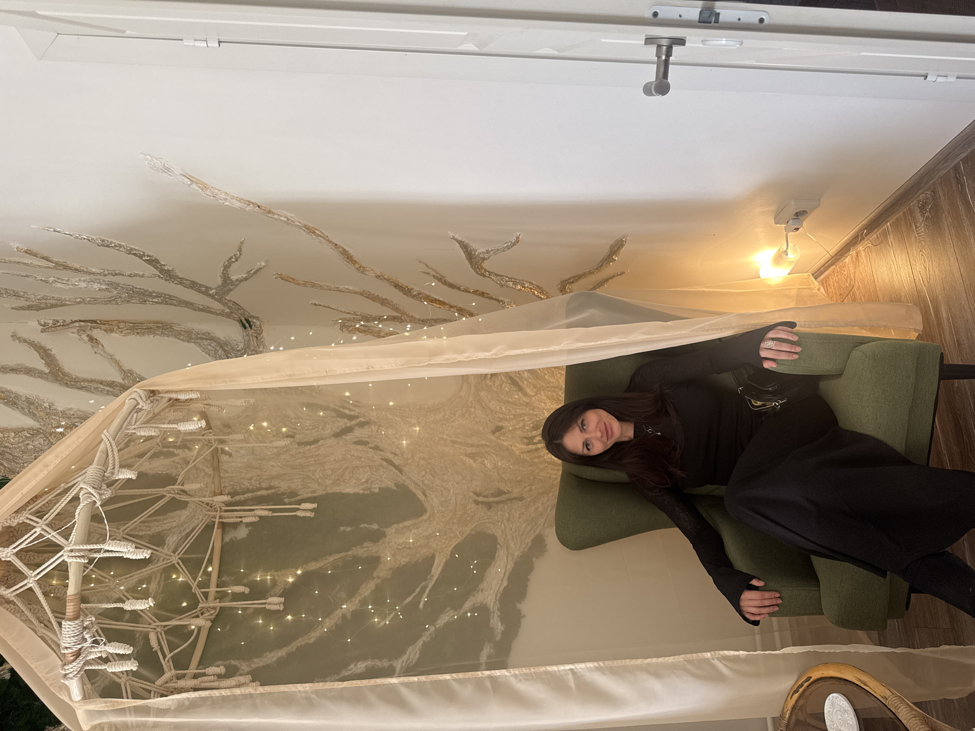 A woman is reclining in an olive-green armchair within an enclosed space with decorative elements reminiscent of a fairy tale. The walls and ceiling are painted with silvery-white tree branch motifs that extend across the surfaces. A canopy structure with a cream-colored fabric cover and white rope embellishments frames the scene. String lights are intertwined within the canopy's framework, adding a soft glow. The room is illuminated by an exposed light bulb fixture on the wall. A partial view of a small round table is visible in the lower right corner. The location appears to be an interior space in an unknown city, but further details are unavailable.