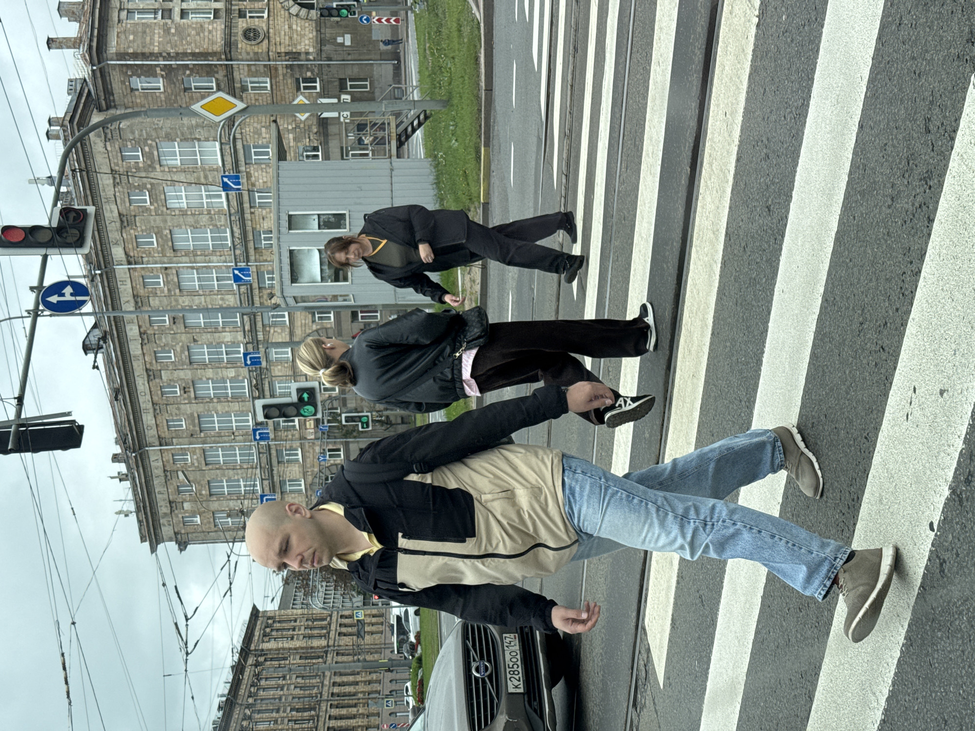 A man with a shaved head, wearing a beige and black jacket, blue jeans, and grey sneakers, walks across a pedestrian crossing. He is on the left side of the frame, walking from left to right. Behind him, two women are also crossing the street. The woman on the left, seen from the back, has blonde hair in a ponytail and wears a black jacket and dark pants. The woman on the right, facing left, has brown hair and wears a black jacket and dark pants. She has a yellow necklace.
The scene is a city street in Saint Petersburg, Russia. The architecture in the background suggests a historic district, with a large, ornate stone building dominating the skyline. Overhead, a complex network of tram wires crisscrosses the sky. Traffic lights and street signs are visible, indicating an urban intersection. A grey car with a license plate that reads "K 28500 142" is parked on the left side of the street. The weather appears overcast. The pedestrian crossing is marked with white stripes on the asphalt.
