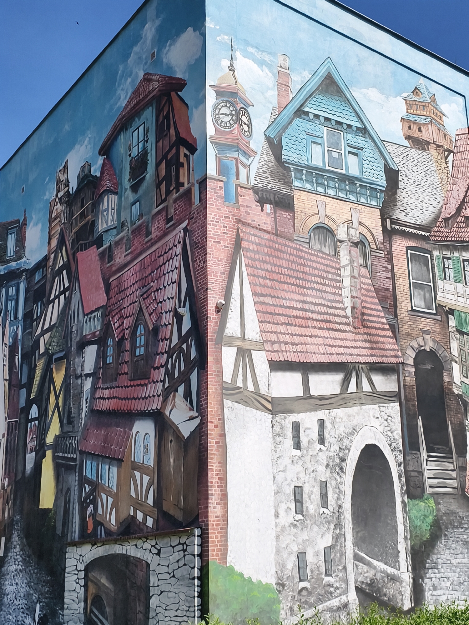 The image shows a mural painted on the side of a building in Tczew, Poland. The mural depicts a street scene with old European-style buildings. The architecture features half-timbered houses, red tiled roofs, and brick facades. There are windows, chimneys, and architectural details like arched doorways and balconies. The sky in the mural is a bright blue with white clouds. The mural wraps around a corner of the building, giving a sense of depth and perspective. There are no people visible in the mural. The scene appears to be a historical or fantasy-inspired urban setting.