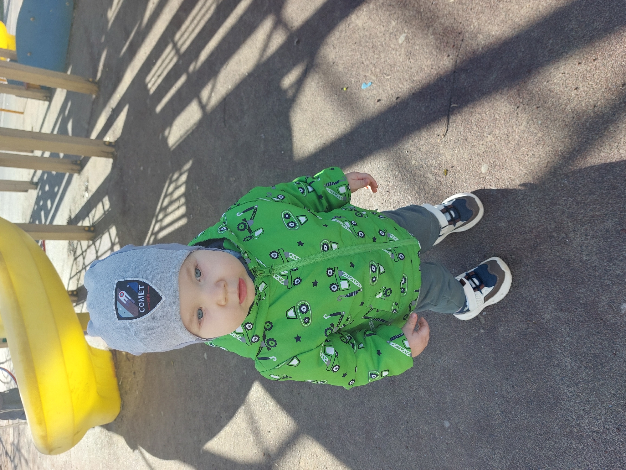 The image features a young child, likely a toddler, standing on a paved surface at an outdoor playground in Chaikovsky, Russia. The child is looking directly at the camera with wide blue eyes and a neutral expression.
The child is dressed in a vibrant green jacket adorned with cartoon-style construction vehicles like excavators, tractors, and stars. They are also wearing grey pants and white, blue, and brown sneakers. A light grey beanie covers the child's head, featuring a patch with the word "COMET" boldly displayed above "SOLO," alongside a graphic of what appears to be a spacecraft or rocket.
Parts of yellow playground equipment, possibly a slide, and wooden support beams are visible on the left side of the frame. The ground, which appears to be asphalt or a similar material, is marked by long, distinct shadows, suggesting a sunny day, likely during the mid-morning or afternoon. The scene depicts a quiet moment at the playground, with the child seemingly pausing from play to interact with the photographer.