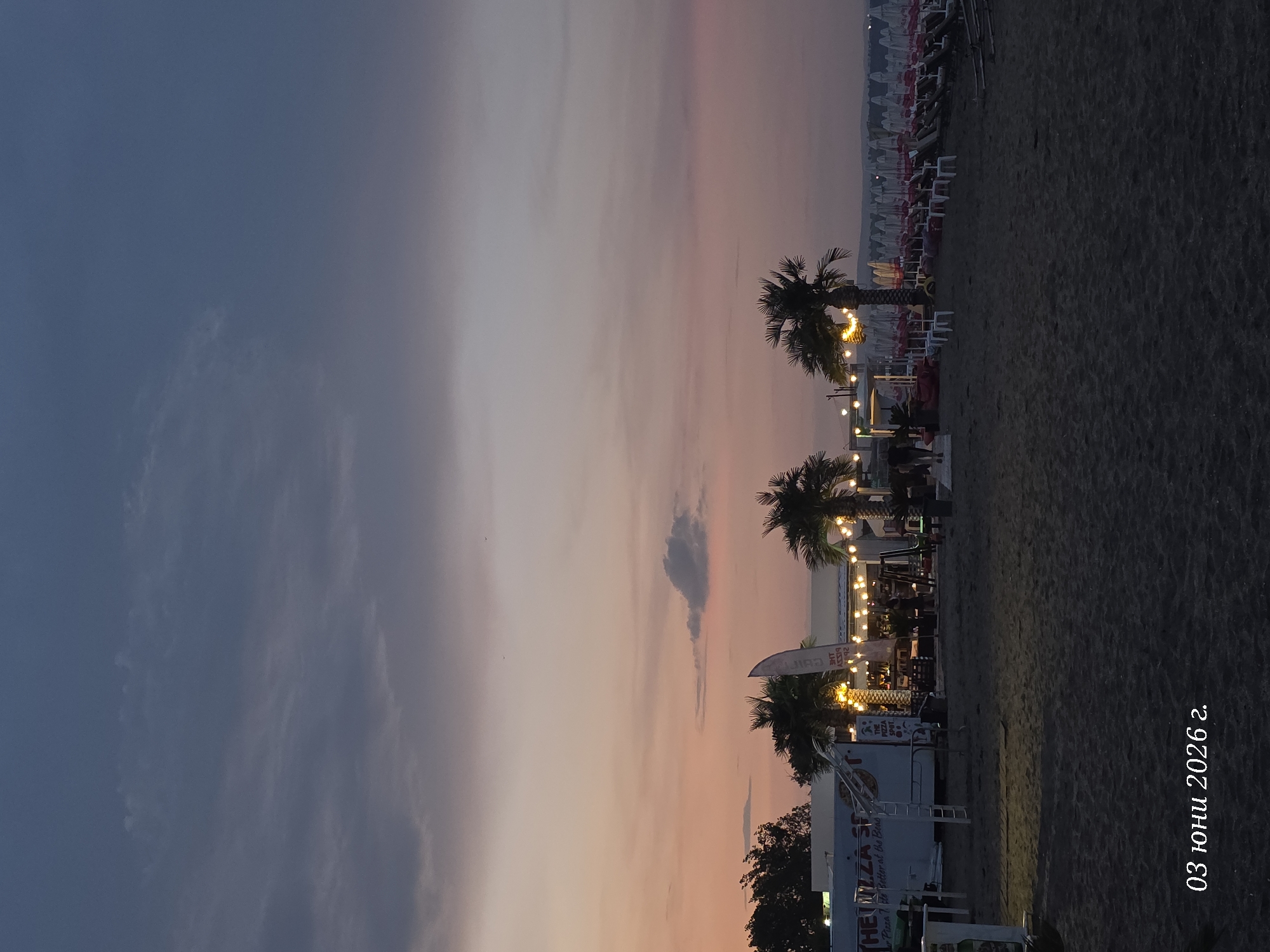 This media file captures a vibrant beach scene at dusk or early night, likely in Burgas, Bulgaria, based on the provided context. The image is taken from a slightly elevated perspective, looking across a sandy beach towards a lighted area.
The sky dominates the upper portion of the frame, displaying a beautiful gradient of colors typical of twilight. It transitions from deep blue on the left to soft purples, pinks, and faint oranges towards the right horizon, with wispy clouds scattered throughout.
In the mid-ground, a row of palm trees adds to the tropical ambiance, silhouetted against the sky but also illuminated by string lights strung between them and various structures. Beneath these lights, there appears to be a lively beachside establishment with tables and chairs. Several indistinct figures can be seen, suggesting people are gathered there, enjoying the evening.
To the right, a white food truck or stand is prominent, featuring the text "THE PIZZA SPOT" and "Pizza & the Better at the Beach." A white flag or banner nearby also displays "PIZZA." Further back and along the shoreline, numerous beach chairs and umbrellas, predominantly white and red, are neatly lined up or stacked, indicating a public or resort beach area. The sandy beach itself occupies the lower right portion of the image.
A notable detail is the date "03 юни 2026 г." (June 3, 2026) written vertically in white on the far right side of the image. The overall atmosphere is relaxed, inviting, and illuminated, suggesting a pleasant evening gathering by the sea.