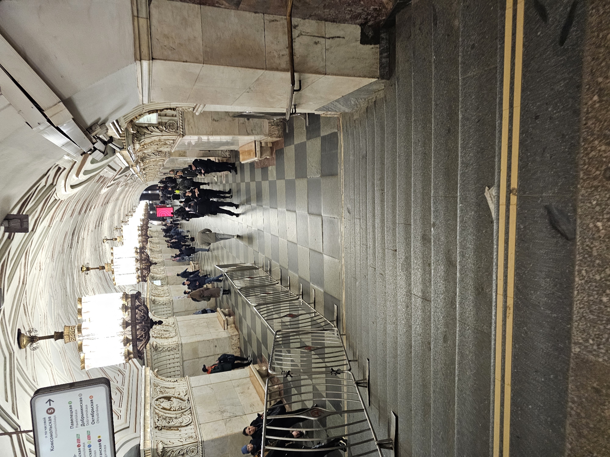 The image depicts a scene within the Moscow Metro system in Russia. The location is the Komsomolskaya station, as indicated by the directional sign displaying the names of connecting stations.
The scene consists of a long perspective view of a corridor with a checkered floor. People are present, standing and walking along the corridor. On the right of the frame are steps with two yellow lines. The walls are decorated with baroque-style moldings and chandeliers. A metal barrier runs alongside the corridor, suggesting an area with pedestrian traffic control.