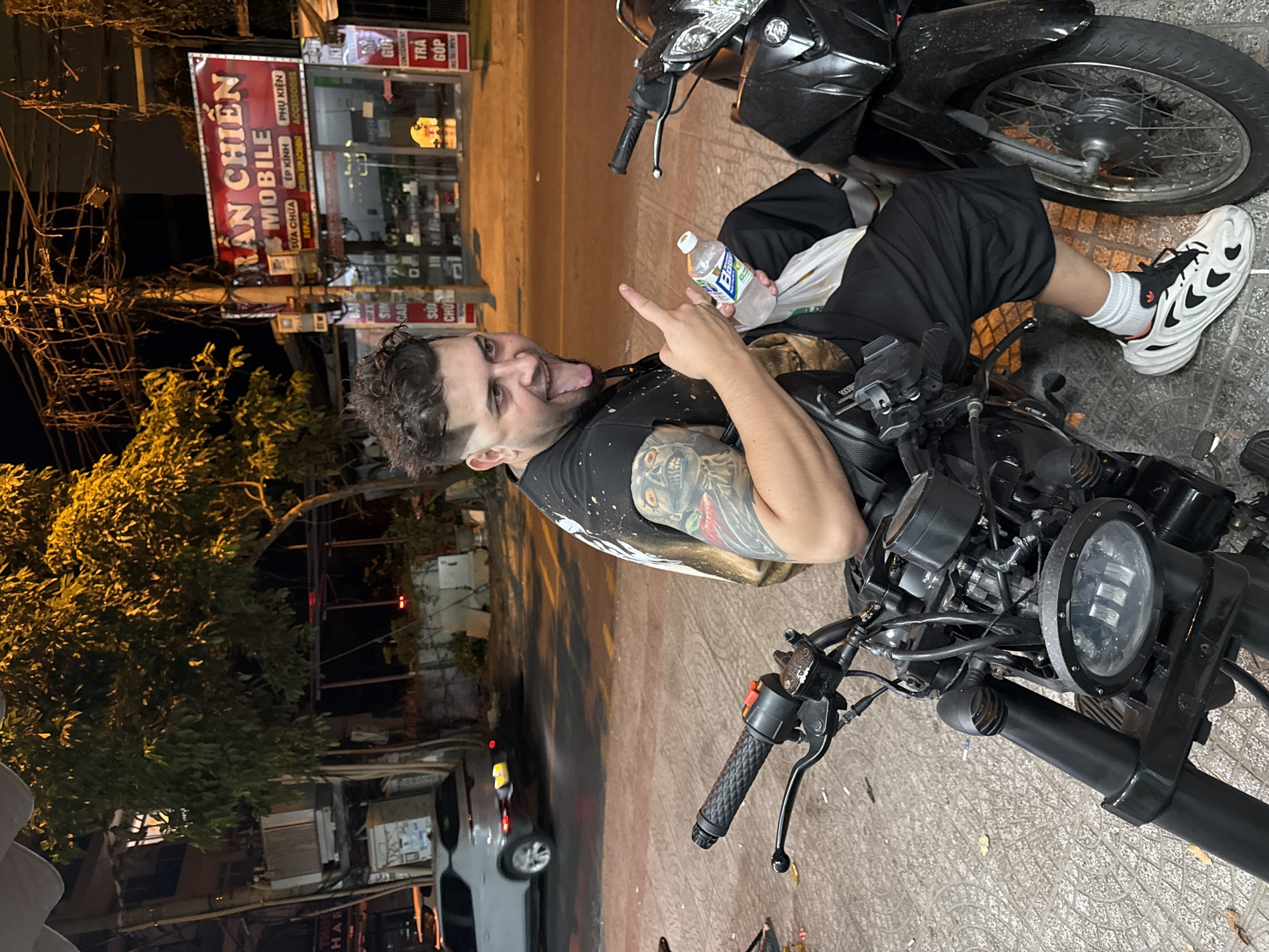 The image depicts a man sitting on a black motorcycle in what appears to be a street in Đà Nẵng, Vietnam. The man is Caucasian, with dark hair and a beard, and has a tattoo on his left arm. He is wearing a black t-shirt and black shorts, and white sneakers. He is holding a bottle of water in his left hand and making a gesture with his right hand. Behind him, there's another motorcycle. Across the street, there's a storefront with a sign that reads "RAN CHIEN MOBILE," offering phone repair services. The street is paved with tile-like textures, and there are streetlights and trees in the background. A car is parked further down the street. The image appears to have been taken at night.