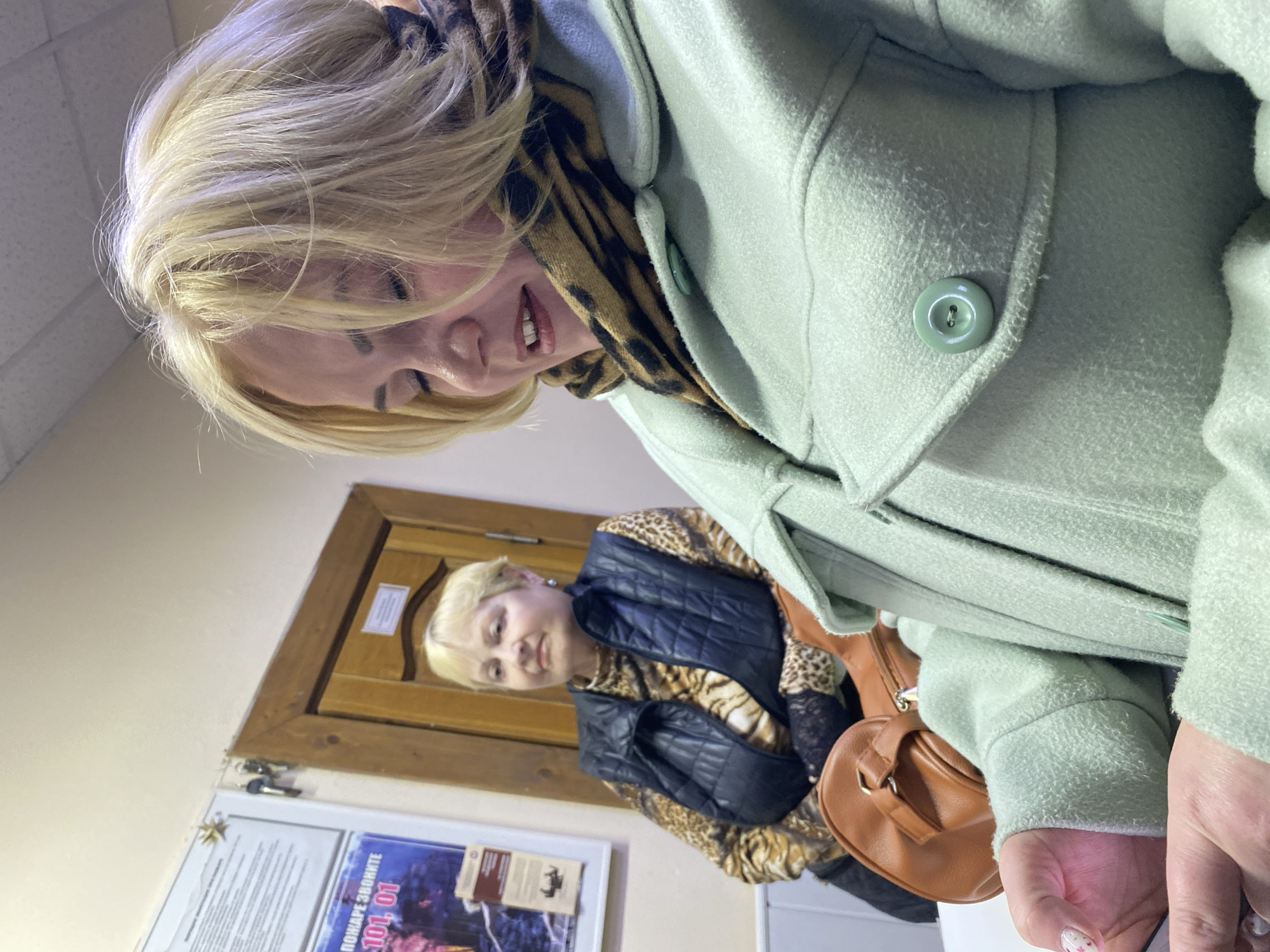 A photograph shows two fair-skinned women in an indoor environment. The primary subject is a blonde woman wearing a light green coat with a round green button. A brown animal-print scarf is visible underneath the coat.
In the background, another blonde woman stands near a wooden door frame. She is wearing a black vest over a brown animal-print shirt and a tan purse. On the wall, a poster can be partially seen, with visible text that reads "ПОЖАРЕ ЗВОНИТЕ 101, 01", likely translated as "Call 101, 01 for a fire." Based on the Cyrillic text, this location could be Noviy, Russia. Additional details such as keys hanging next to the door can be seen.