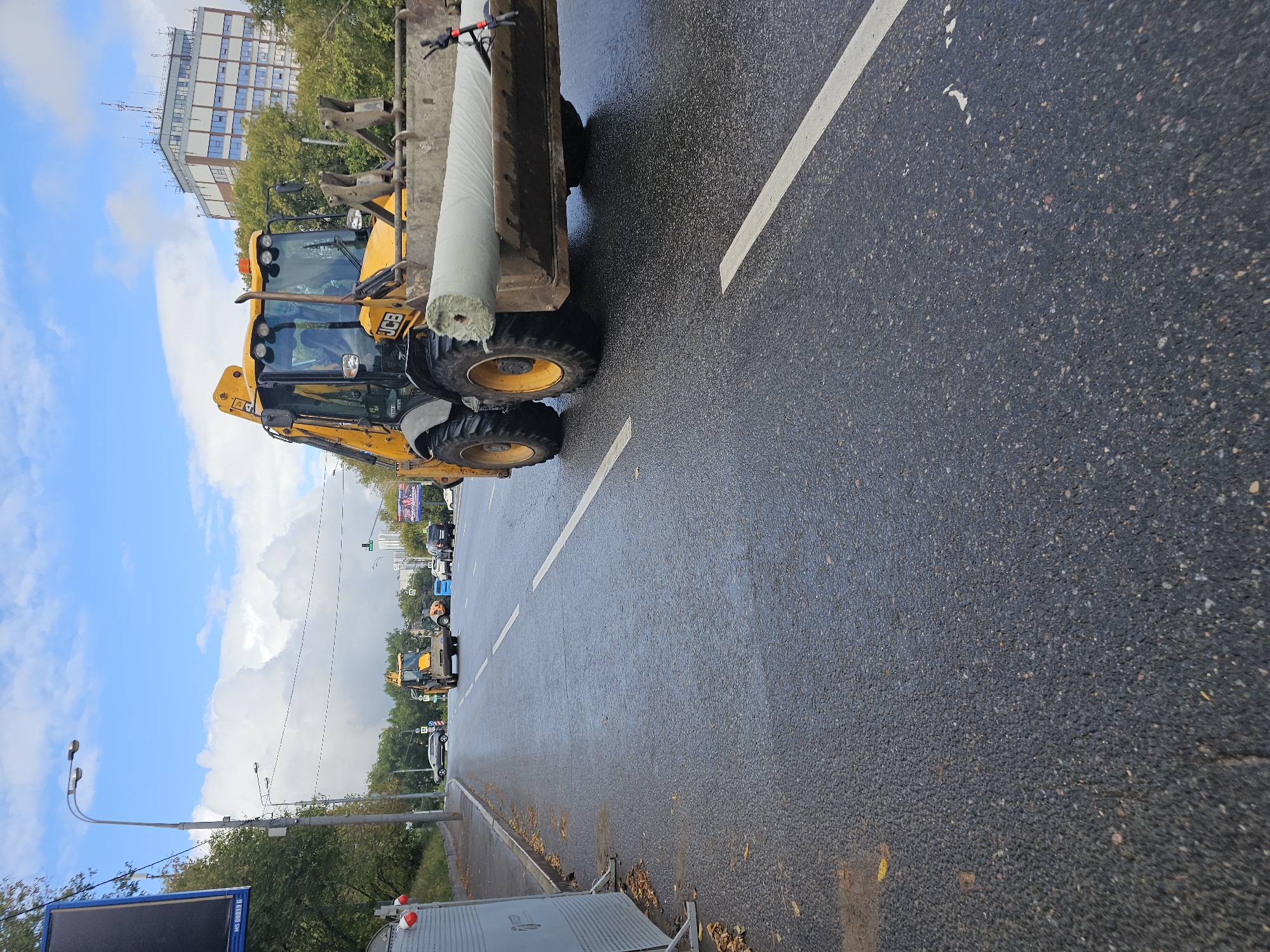 In Moscow, Russia, on a cloudy day, a yellow JCB backhoe loader is driving on a wet asphalt road, carrying a large roll of light-colored material in its front loader. The backhoe has the "JCB" logo visible on its side. The road has a white line dividing the lanes. To the left of the road, there is a sidewalk with some fallen leaves and a temporary barrier with red and white markers. In the background, there are trees, other vehicles including a smaller orange construction vehicle, and a multi-story building. The sky is blue with large, puffy white clouds.