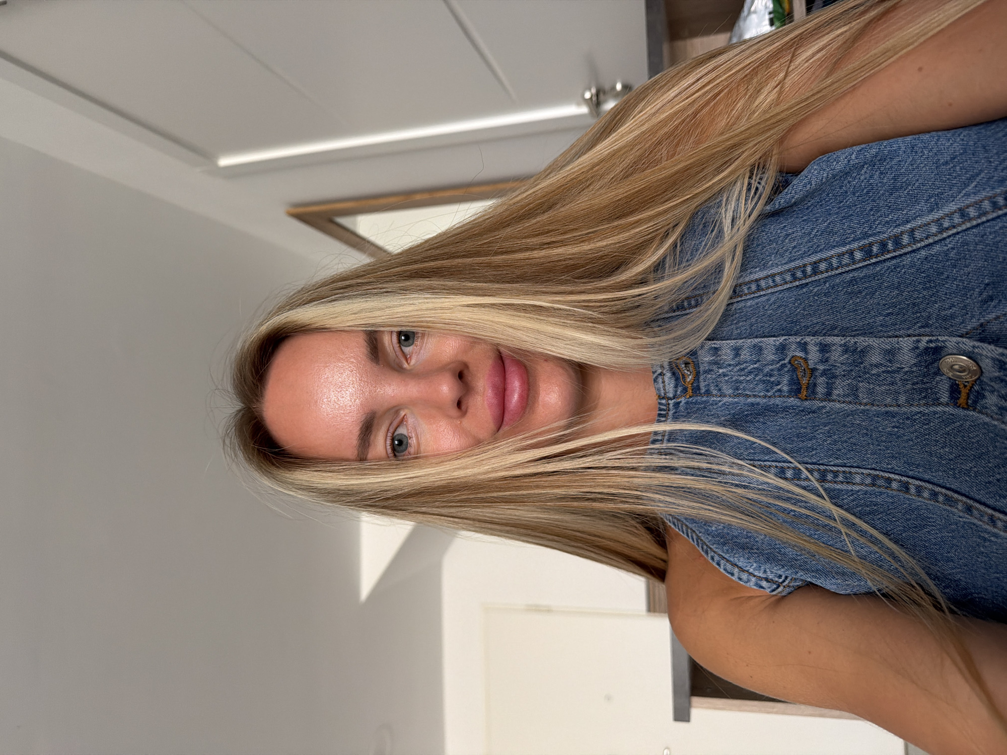A medium shot captures a fair-skinned woman with blonde hair and blue eyes, facing forward. She is wearing a denim dress and her skin has a slightly rosy hue, suggesting it might be sun-kissed. The background is a softly lit interior with a wall and a doorway visible, hinting at a domestic setting within Finestrat, Spain. The lighting appears to be natural, possibly from a nearby window, casting a gentle illumination on the scene.