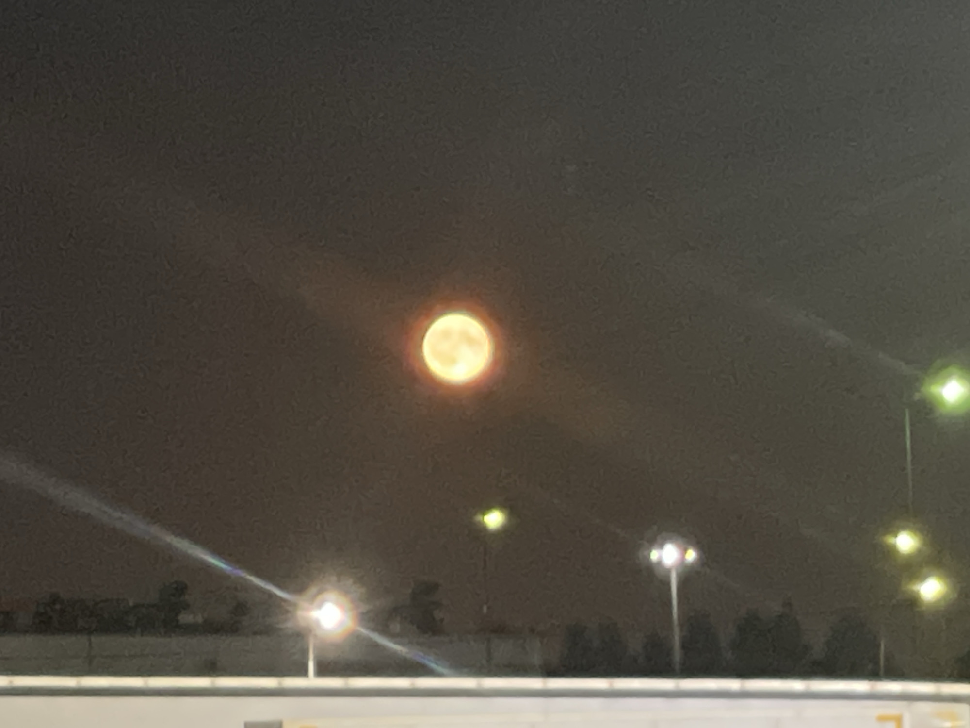 The image shows a full moon, illuminated with a warm, orange hue, against a dark, hazy sky. The moon appears to be low in the sky, possibly near the horizon. In the foreground, a faint, blurred line of lights stretches across the bottom of the frame, suggesting a road or cityscape at night. There are also several other lights in the middle ground, some appearing as bright orbs and others as streaks of light, possibly from streetlights or vehicles. The overall impression is one of a nocturnal scene in a semi-urban or rural environment, with the moon being the dominant celestial feature. There is no discernible information about the specific location in the city of Veshki, Russia.