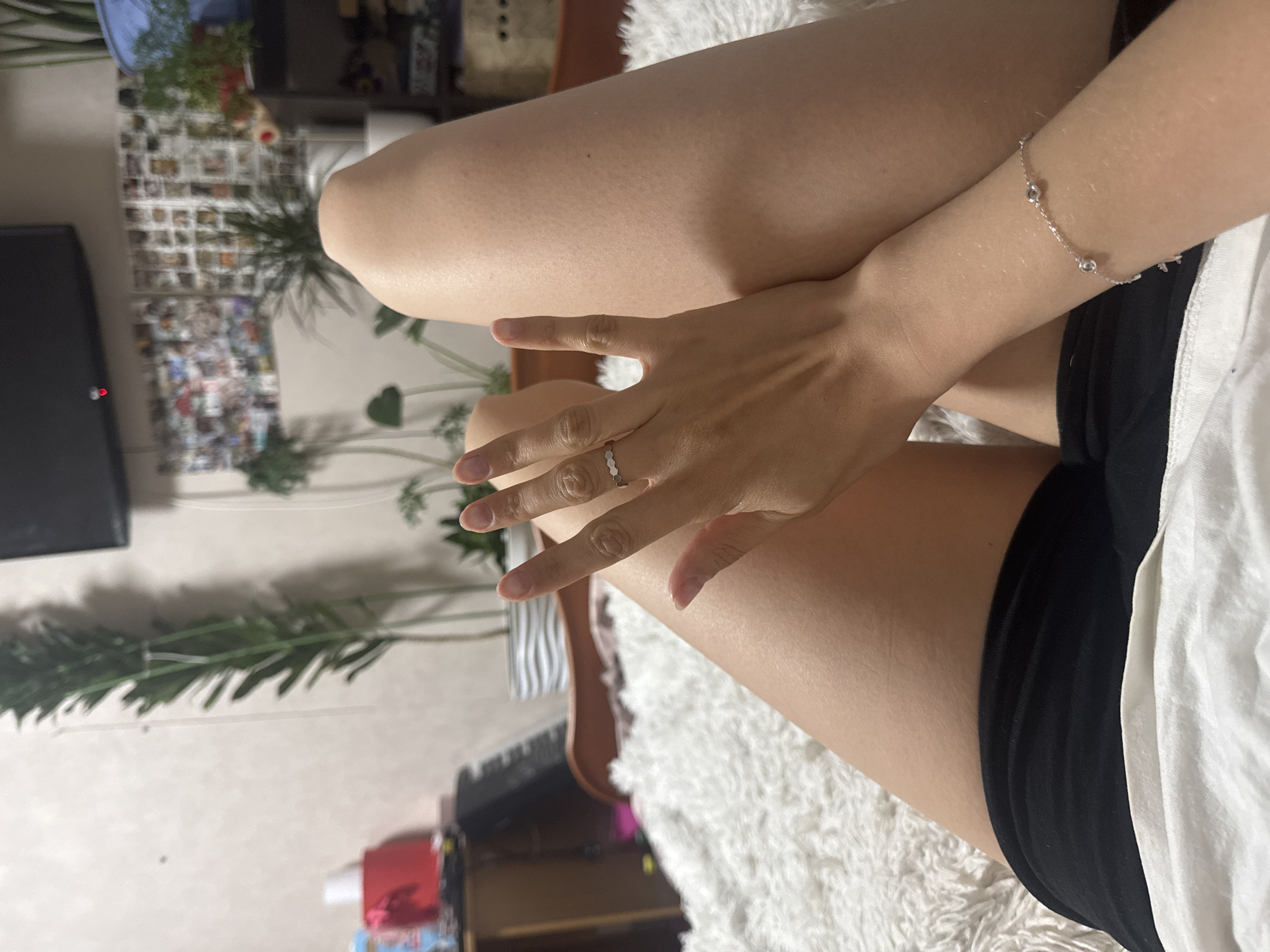 The image displays a partial, close-up view of a person relaxing indoors. The perspective is from above, looking down at the individual's legs, left arm, and hand. The image is rotated 90 degrees clockwise, with the person's body extending diagonally across the frame from bottom-left to top-right.
The central subjects are the person's bare legs, which are bent and casually positioned, and their left arm with the hand resting on a thigh. A silver ring with a distinct patterned band (possibly hexagonal or scalloped) is visible on the ring finger of the left hand. A delicate silver bracelet featuring three small, round, reflective charms adorns the left wrist. The person is wearing black shorts, with a piece of white fabric—likely a shirt or a blanket—visible beside their hip. Their legs are resting on a plush, white, fluffy surface, which appears to be a rug or a blanket.
The background reveals an indoor setting, likely a living room or bedroom. On the far left, a dark television screen with a small red indicator light is partially visible. A wall behind the person is decorated with an extensive grid of numerous small photographs or postcards. Various green houseplants, both large and small, are scattered throughout the scene, contributing to a cozy atmosphere. The lighting is soft and ambient. No discernible text is visible anywhere in the image. The scene suggests a moment of calm relaxation.