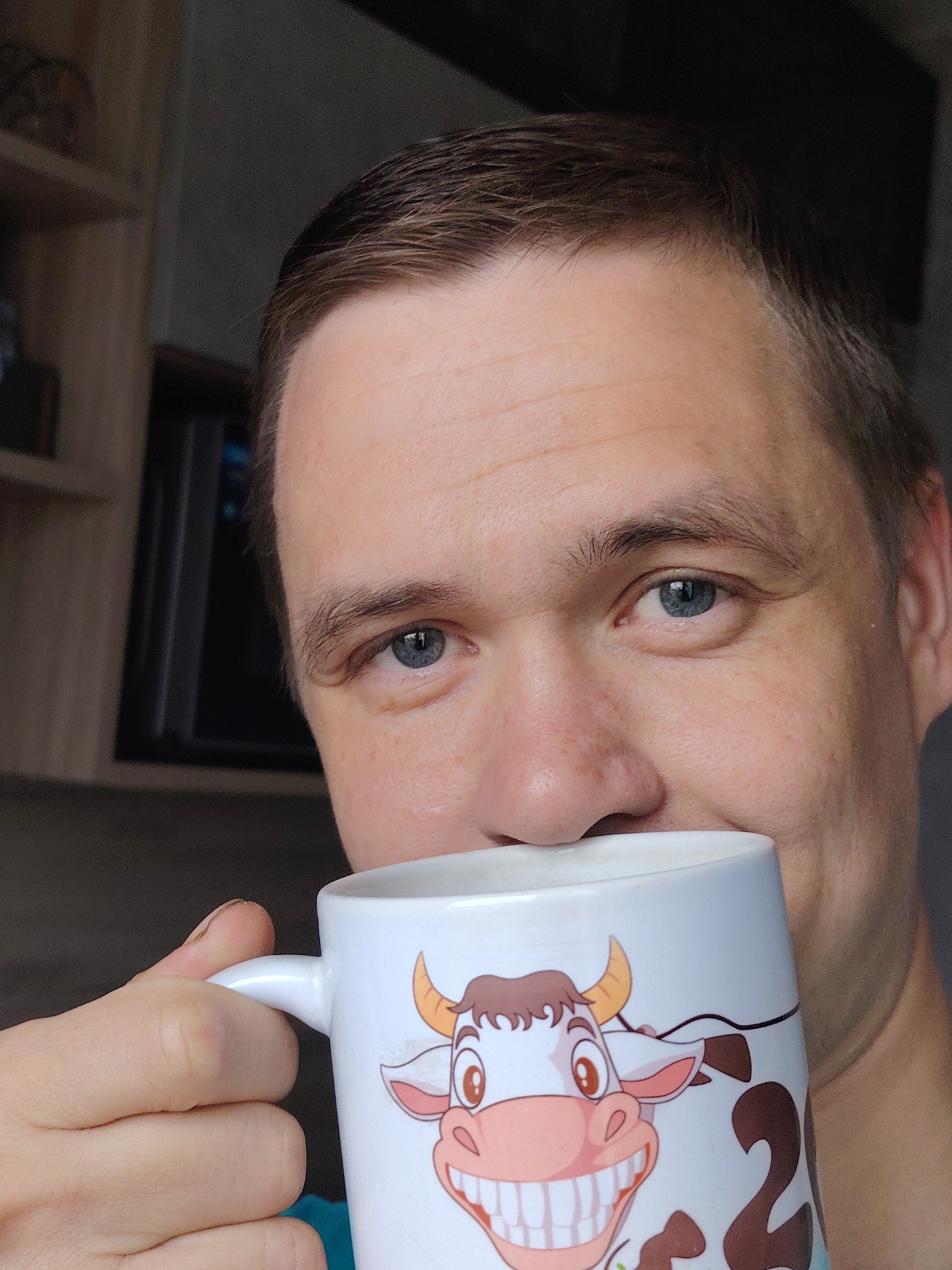 A man is holding a white mug with a cartoon cow on it. The man has short brown hair, blue eyes, and fair skin. He is partially obscured by the mug, which is held close to his face. The background includes a dark gray wall and a wooden cabinet with a black electronic device inside. The image was most likely taken indoors. The location of the image within the city of Chaikovsky, Russia cannot be determined from the image alone.