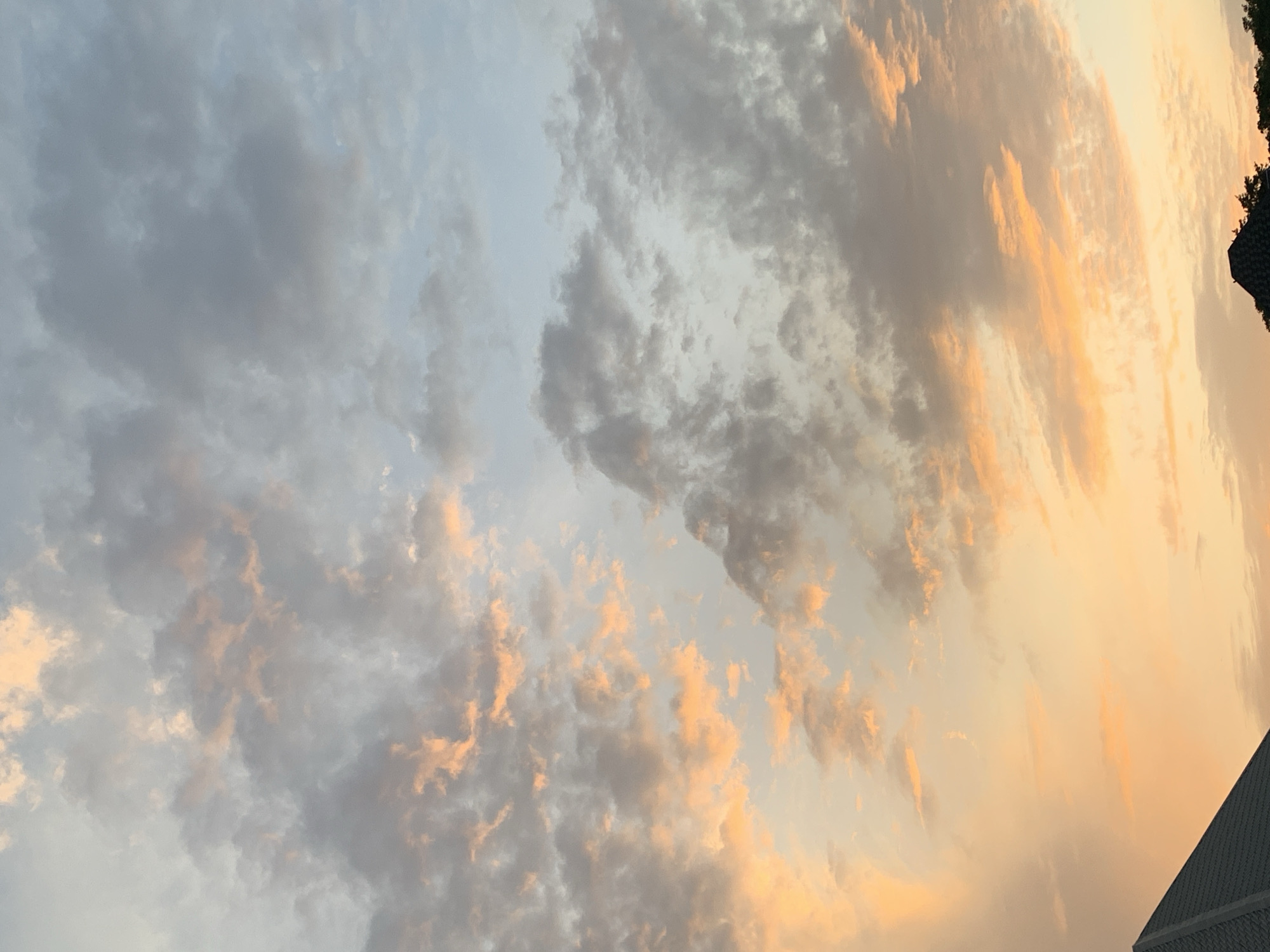 The image depicts a sky filled with clouds at sunset. The clouds are various shades of gray and white, with orange and yellow hues from the setting sun illuminating their edges. The sky transitions from a lighter blue-gray on the left to a warm orange-yellow on the right. A small section of dark green foliage and a portion of a gray building are visible in the lower right corner. The location is New Stadium, Nigeria. The specific details of people, actions, and interactions are not detectable.