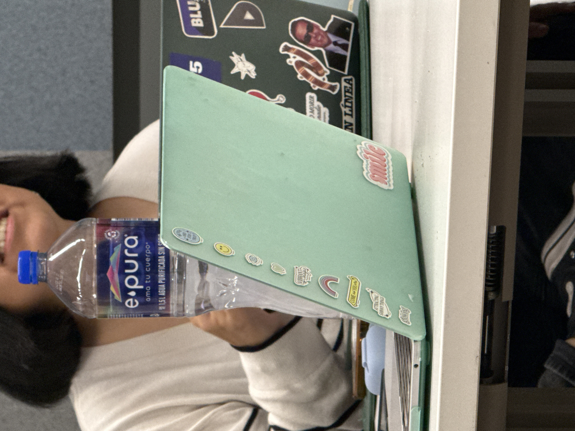 The image, rotated 90 degrees clockwise, captures an indoor scene, likely at a desk or table. A person, whose head, shoulders, and arm are visible on the left side, appears to be smiling or laughing. Their dark hair is prominent, and they are wearing a light-colored top with dark striped cuffs.
In the foreground, a light green laptop is open, angled towards the viewer. Its lid is adorned with several small, colorful stickers, including text that reads "smile," "VIBRANT," "COME YOU are," "ATTRACTING SHINNY THINGS," and "glowing." Other stickers include a happy face, an eye, a heart, and a rainbow. A clear plastic water bottle, positioned upright from the perspective of the original photo, is placed near the laptop. The bottle is from the brand "e.pura" and its label states "ama tu cuerpo" (love your body) and "1.5L AGUA PURIFICADA SIN SORI."
Behind the light green laptop, another device, likely another laptop, is visible. It is dark-colored and also covered in various stickers. Prominent stickers on this device include "BLU," the number "5," and text "IN LÍNEA." There's also a sticker featuring a man's face, possibly a political figure, wearing sunglasses and a suit. Other stickers include a stylized "A," a star, a scarf, and text "MORIR nadie."
The background shows a blue-grey textured wall or partition on the left, and a light-colored vertical surface, possibly a desk leg or part of a shelf, on the right. The overall impression is one of a casual, personalized workspace.