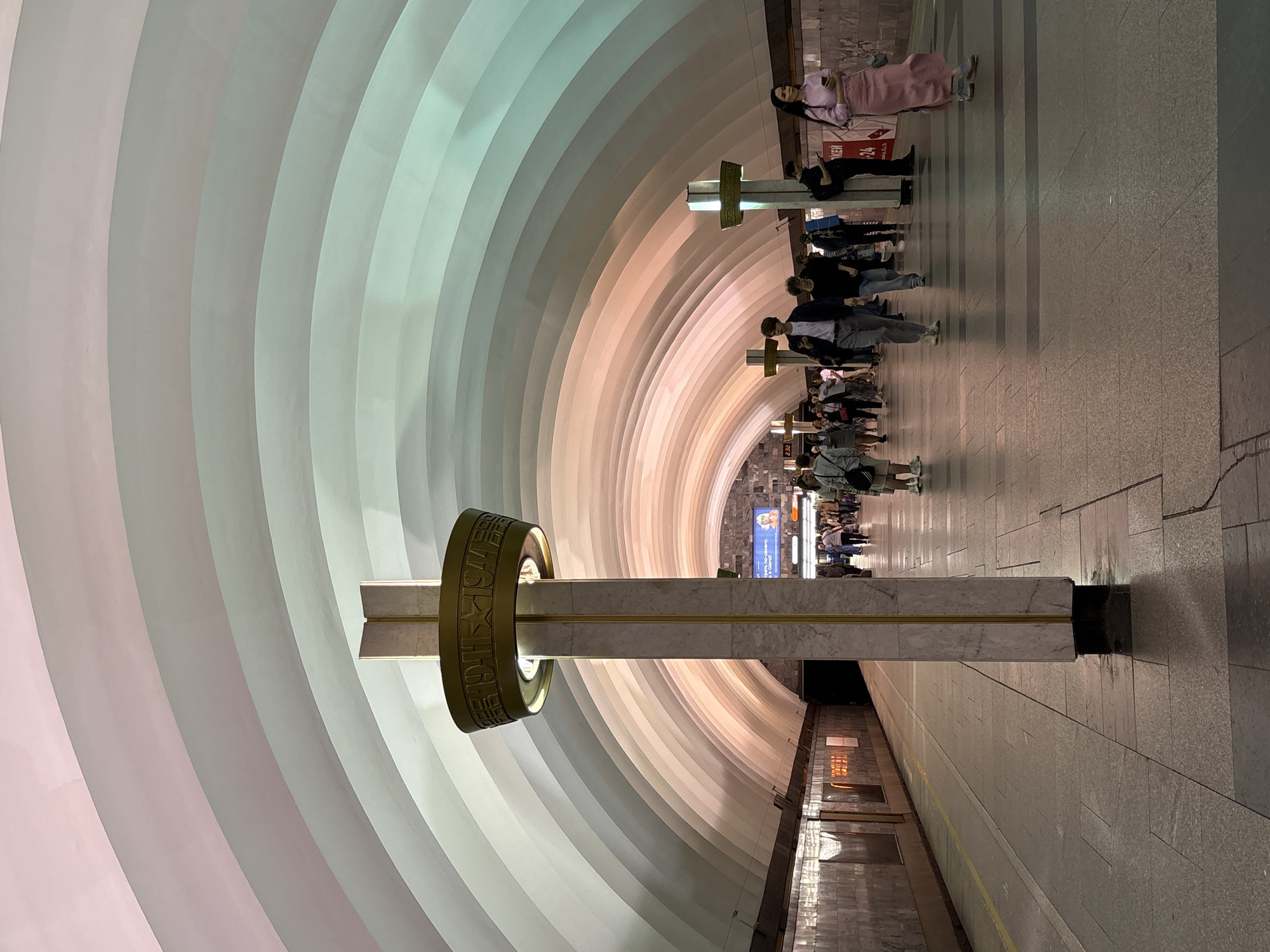 This image shows a view of the platform of Narvskaya metro station in Saint Petersburg, Russia. The station has a distinctive arched ceiling that is illuminated with bands of pastel colors, transitioning from pinks to greens. A tall, marble-clad pillar with gold accents stands in the foreground, topped with a circular golden fixture decorated with inscriptions and symbols. People are walking along the platform, some are looking at their phones, and others are walking together. The platform is paved with granite tiles. The walls of the station are lined with marble. A digital display is visible on the wall in the background.