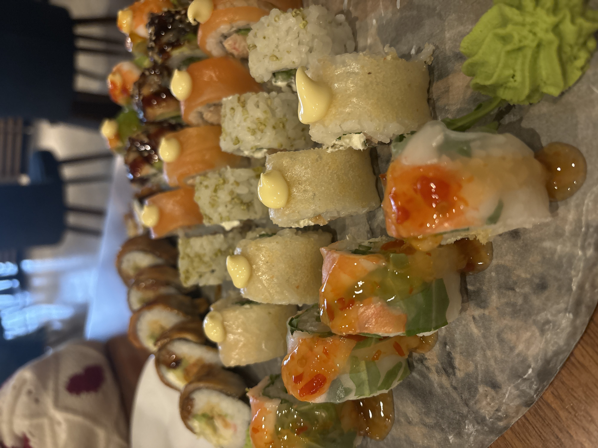 A close-up shot of a sushi platter on a wooden table. The platter contains various types of sushi rolls, including those topped with mayonnaise-like sauce, some covered in seeds, and spring rolls with what appears to be a sweet and spicy sauce drizzled on top. A dollop of wasabi is visible on the right side of the platter. The lighting is soft, and the focus is on the sushi. The scene does not definitively place the location in Varna, Bulgaria; there is no clear visual indication of the city. There are no people clearly visible in the image.