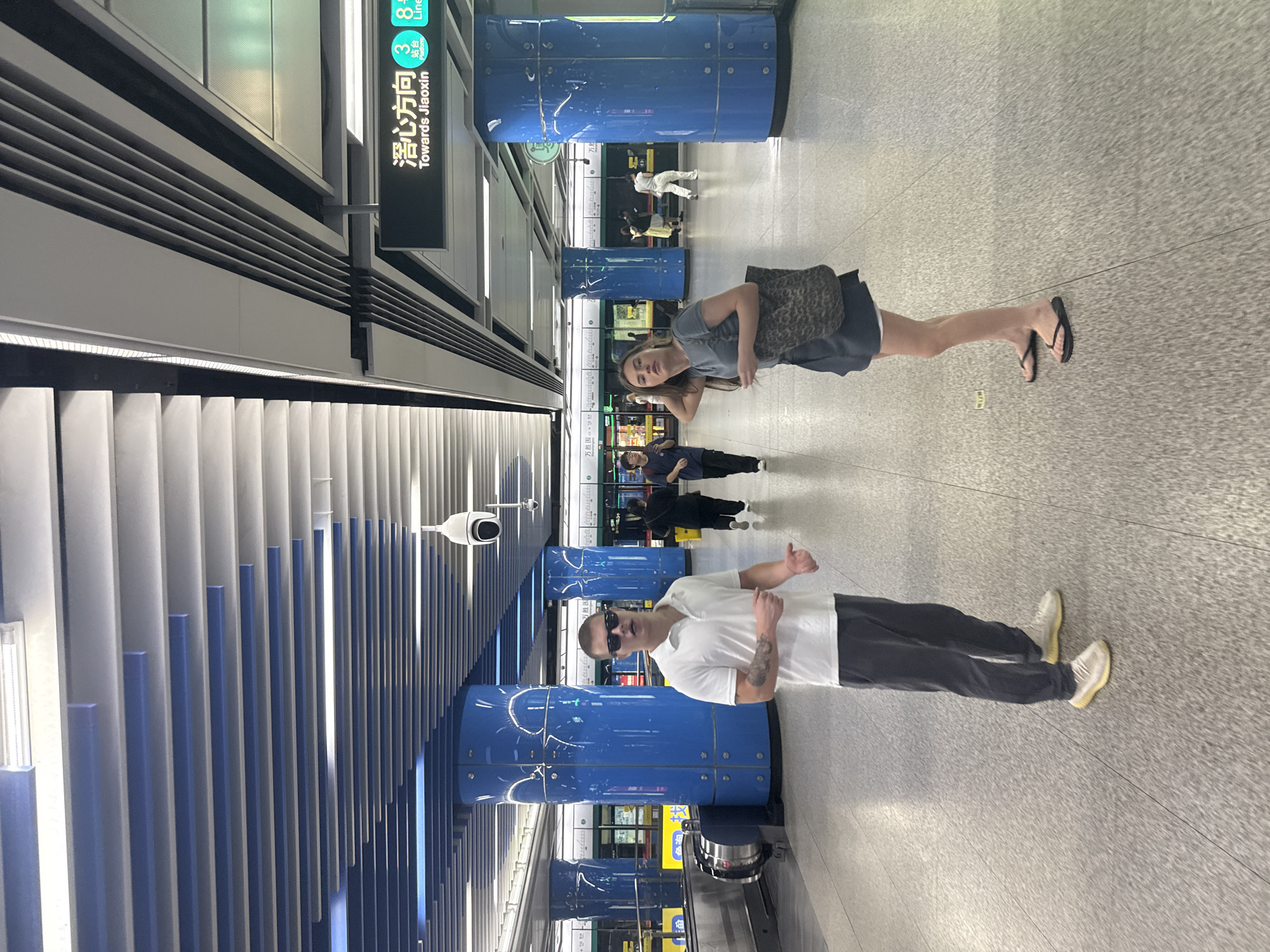 In Haizhu District, China, a man and a woman stand on the concourse of a modern subway station. The man, wearing sunglasses, a white polo shirt, and dark pants, is positioned slightly left of the frame's center, gesturing with his right hand. He is wearing yellow sneakers. The woman, to his right, is wearing a dark grey t-shirt dress and black flip-flops, carrying a leopard-print tote bag over her shoulder and adjusting her hair. In the background, blue cylindrical columns punctuate the space, and a subway train is visible at the platform. Overhead, a segmented white and blue ceiling features lighting fixtures and security cameras. A prominent sign in Chinese and English indicates "Towards Jiaoxin, Platform 3, Line 8". The floor is a speckled grey tile. Several other individuals are present, walking in the background.