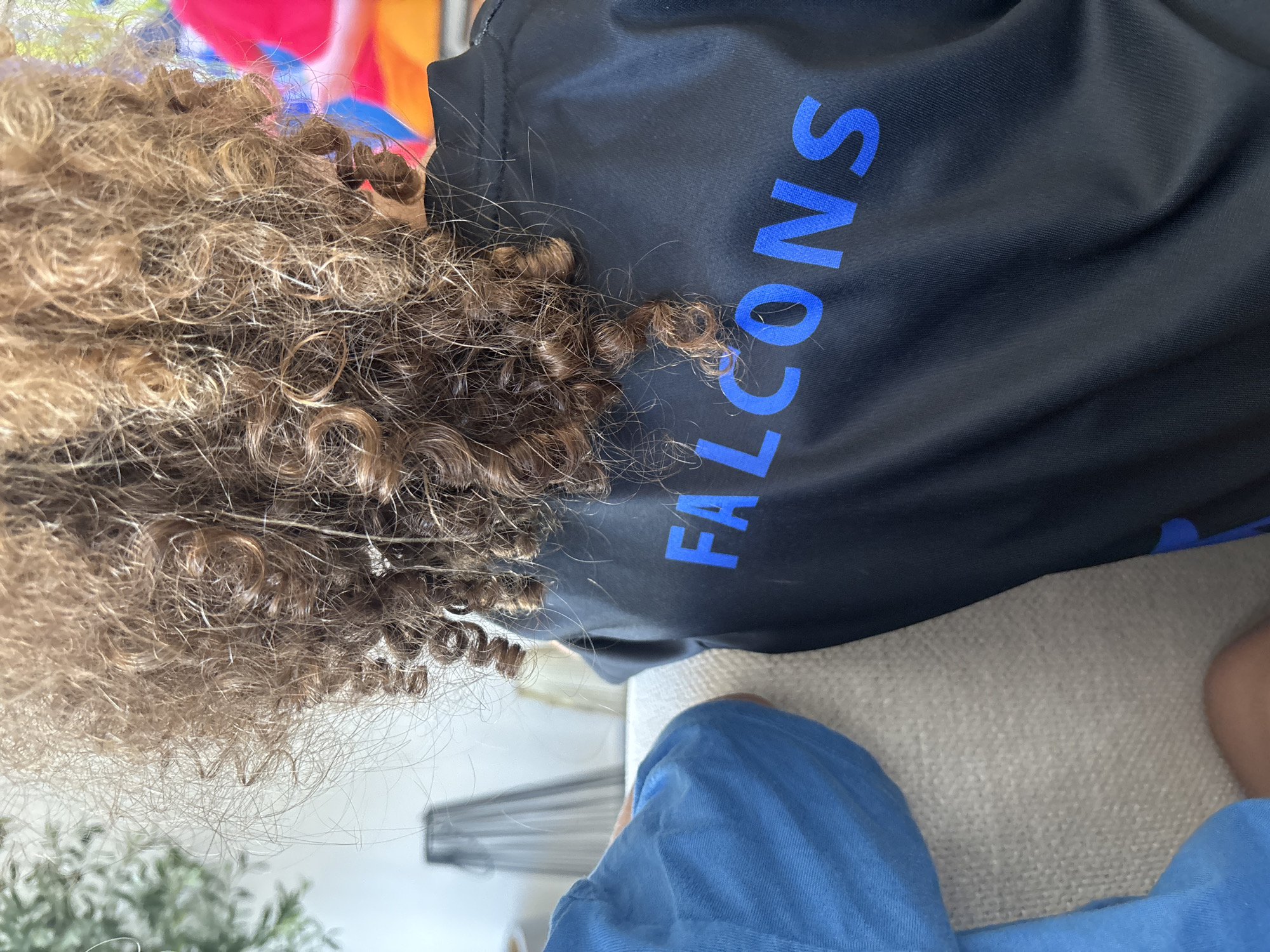 A close-up, rear-view shot shows the back of a person's head and shoulders, with curly brown hair obscuring part of their face. They are wearing a black shirt with the word "FALCONS" written in large, bright blue, block letters across the back. The shirt appears to be made of a synthetic material, possibly athletic wear. The person is seated on a textured, light-colored couch, next to another person wearing a blue shirt. The background is softly blurred, suggesting an indoor setting. To the left, a corner of a modern lamp or decorative object is visible, and further back, a large, colorful piece of abstract art or a screen display with vibrant hues of pink, blue, and yellow is discernible. The lighting is bright and appears to be natural, casting soft shadows. The overall ambiance is casual and relaxed, likely within a home or a lounge area.