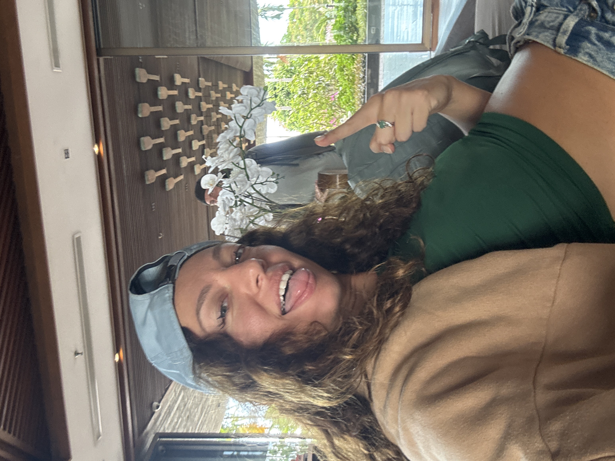 A woman with curly brown hair is sticking out her tongue in a selfie. She is wearing a light blue baseball cap, a tan shirt, and a green top. She is also wearing a ring on her finger.
In the background, there are decorations and objects in the room. Behind her is a wall with a repeating pattern of small, light-colored, bottle-shaped objects. There is a large flower arrangement with white orchids in front of a mirror reflecting trees and greenery. The reflection includes other people in the room. The room appears to be indoors, potentially in a hotel or spa, but the image does not confirm its location in Đà Nẵng, Vietnam.