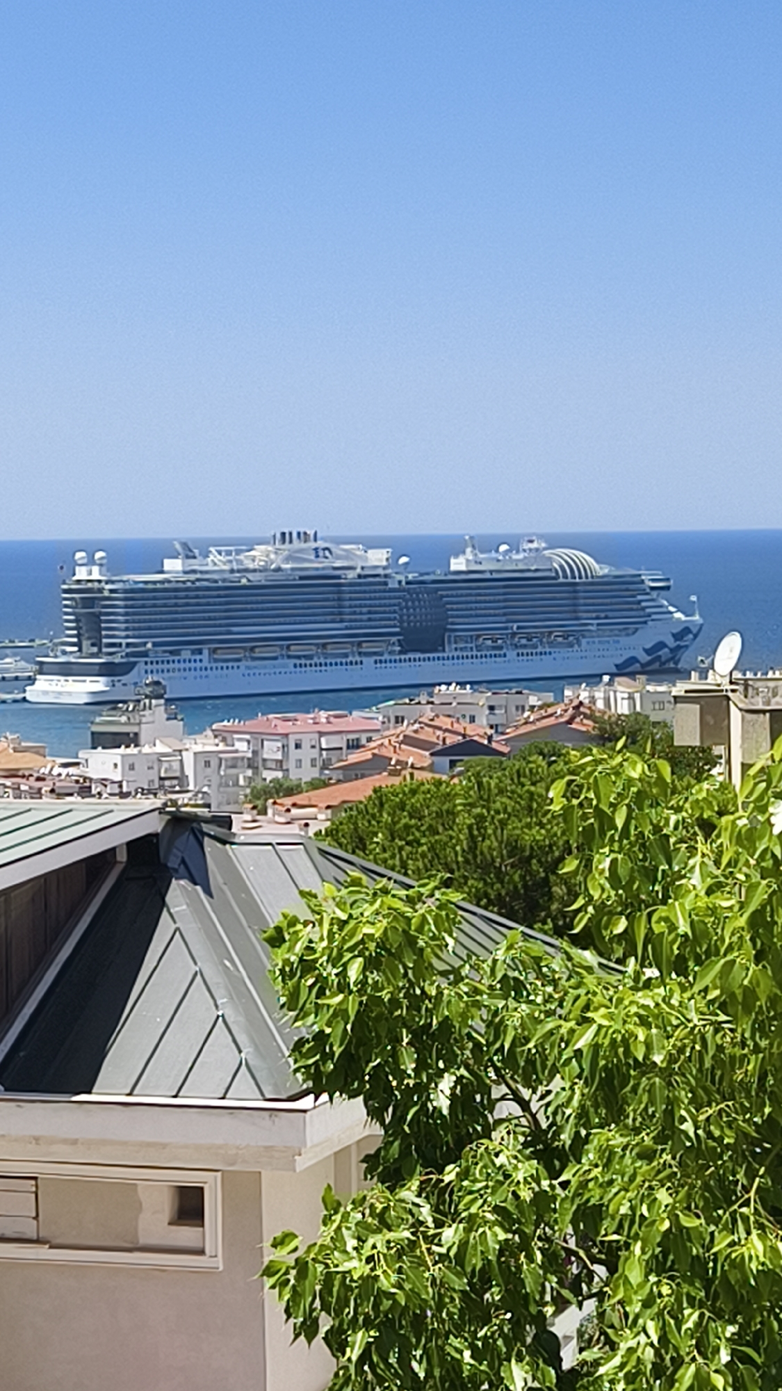 An aerial perspective captures a vast cruise ship docked in the blue waters off the coast of Kuşadası, Turkey, on a clear, sunny day. The ship, predominantly white and blue with multiple decks, is positioned in the distance. In the foreground, the rooftops of Kuşadası are visible, characterized by a mix of white buildings with red-tiled roofs and some modern structures. Lush green trees and foliage partially obscure the view of the city, suggesting a slightly elevated vantage point from a hillside or a building. The sky is a cloudless, vibrant blue.