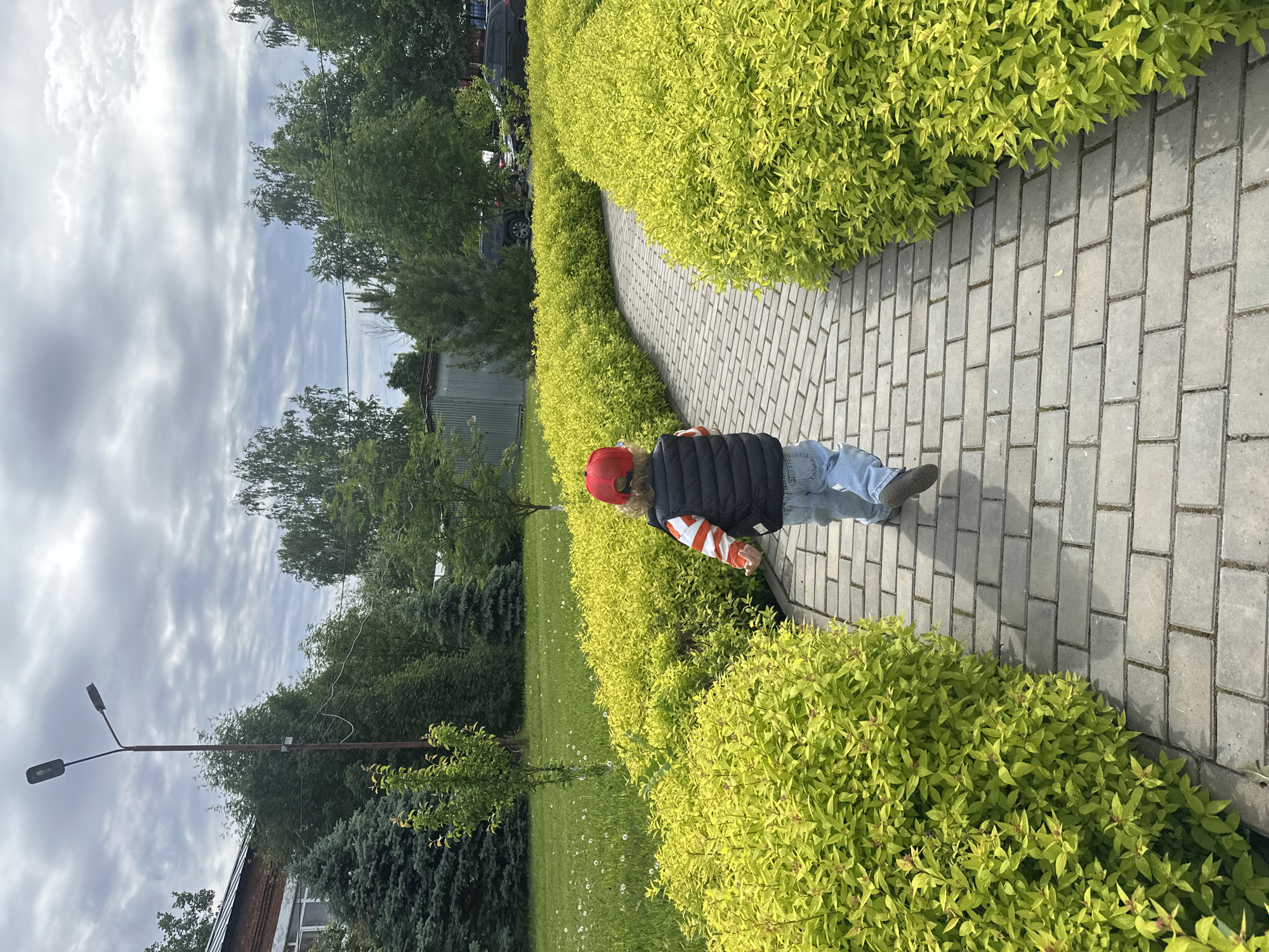 A child with a red cap, a dark vest, a striped orange and white shirt, and blue jeans walks on a gray brick path surrounded by green bushes in a park. The location is Votkinsk, Russia. The sky is overcast. The child appears to be walking away from the camera. The path is curving slightly. In the background, there are trees and a small building. A lamppost is visible to the left. There are parked vehicles in the far background. The ground is grassy with small white flowers scattered throughout.