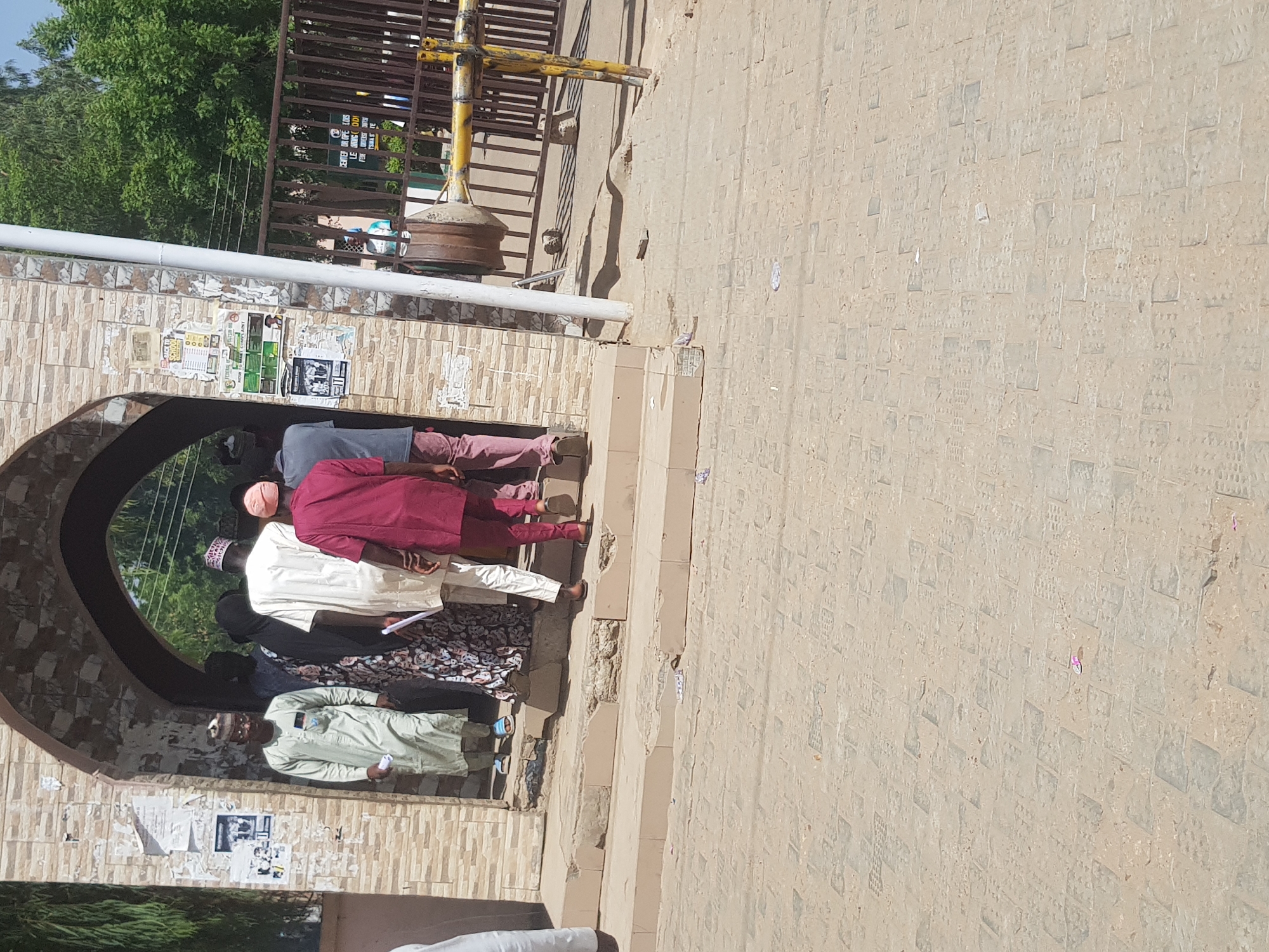 The image shows a group of people standing near an arched entranceway in Dabawa, Nigeria. The entranceway is constructed of tan bricks, with peeling posters affixed to its walls. A metal structure is visible above the archway. The people are dressed in various styles of traditional Nigerian clothing, including gowns and head coverings. The ground is paved with stone blocks.