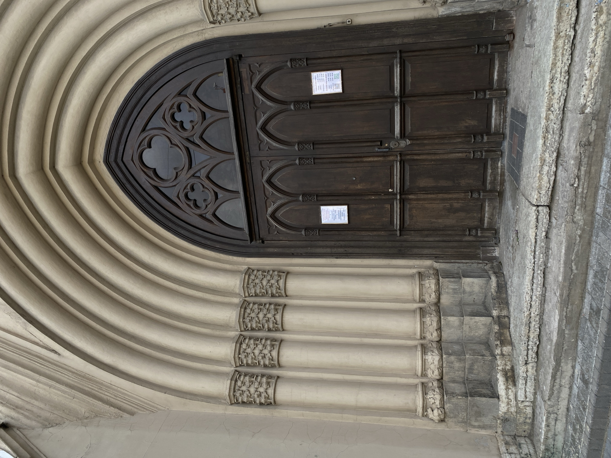 A closed, arched, dark-brown wooden door with decorative quatrefoil glass panels, set in a beige stone archway in Saint Petersburg, Russia. The archway features layered concentric arches with decorative columns on the sides. Two white information notices are affixed to the upper portion of the door. The surrounding stonework exhibits signs of weathering and age.