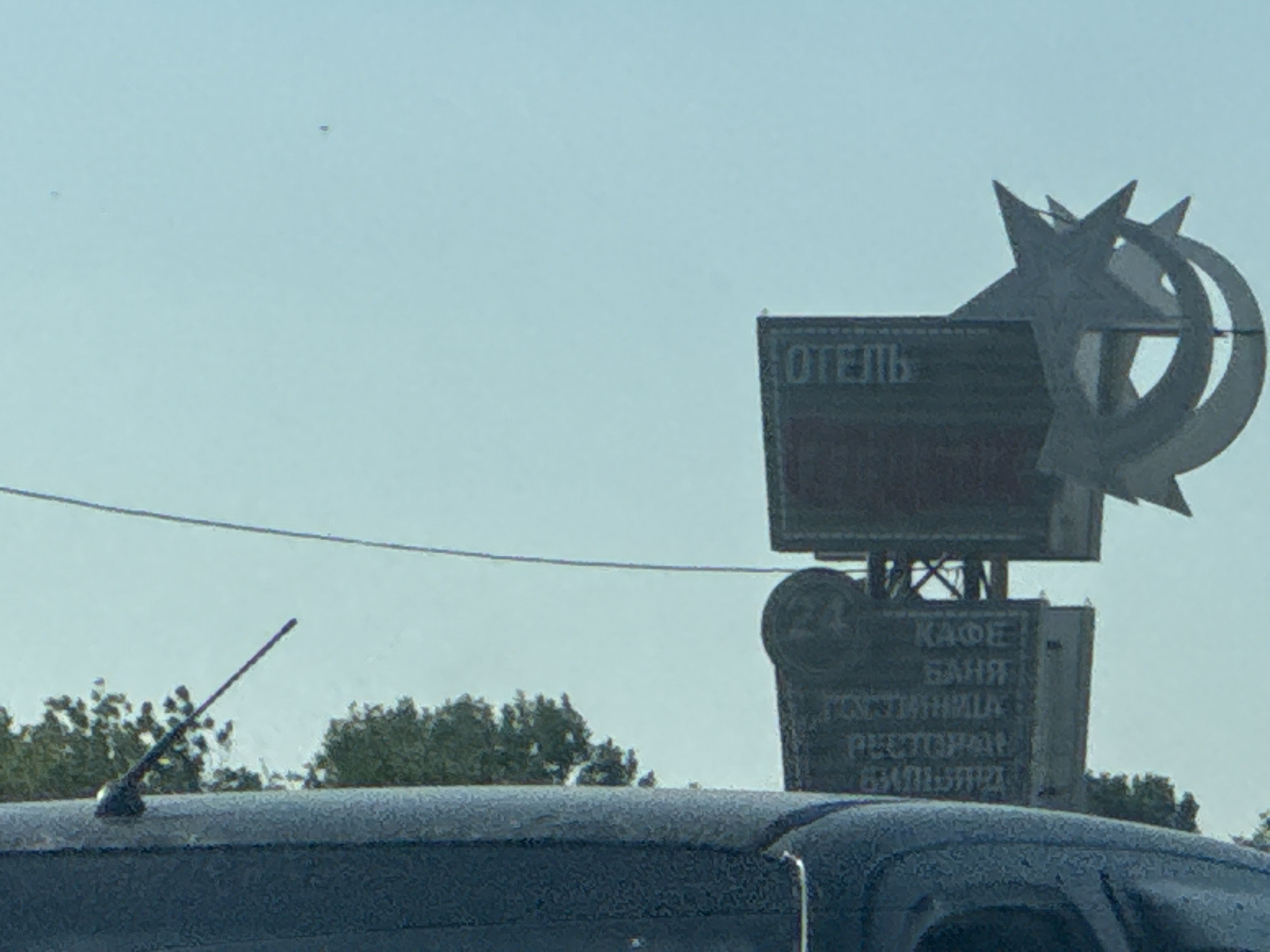 A signpost with an "ОТЕЛЬ" (Hotel) sign and a star and crescent moon emblem is visible against a pale blue sky. Below the hotel sign, another sign lists services: "КАФЕ" (Cafe), "БАНЯ" (Banya/Sauna), "ГОСТИНИЦА" (Hotel/Inn), "РЕСТОРАН" (Restaurant), and "БИЛЬЯРД" (Billiards). The signpost is situated near trees and a car's roof antenna. The image appears to be taken from within a vehicle. The provided location is Perevolotsky, Russia.