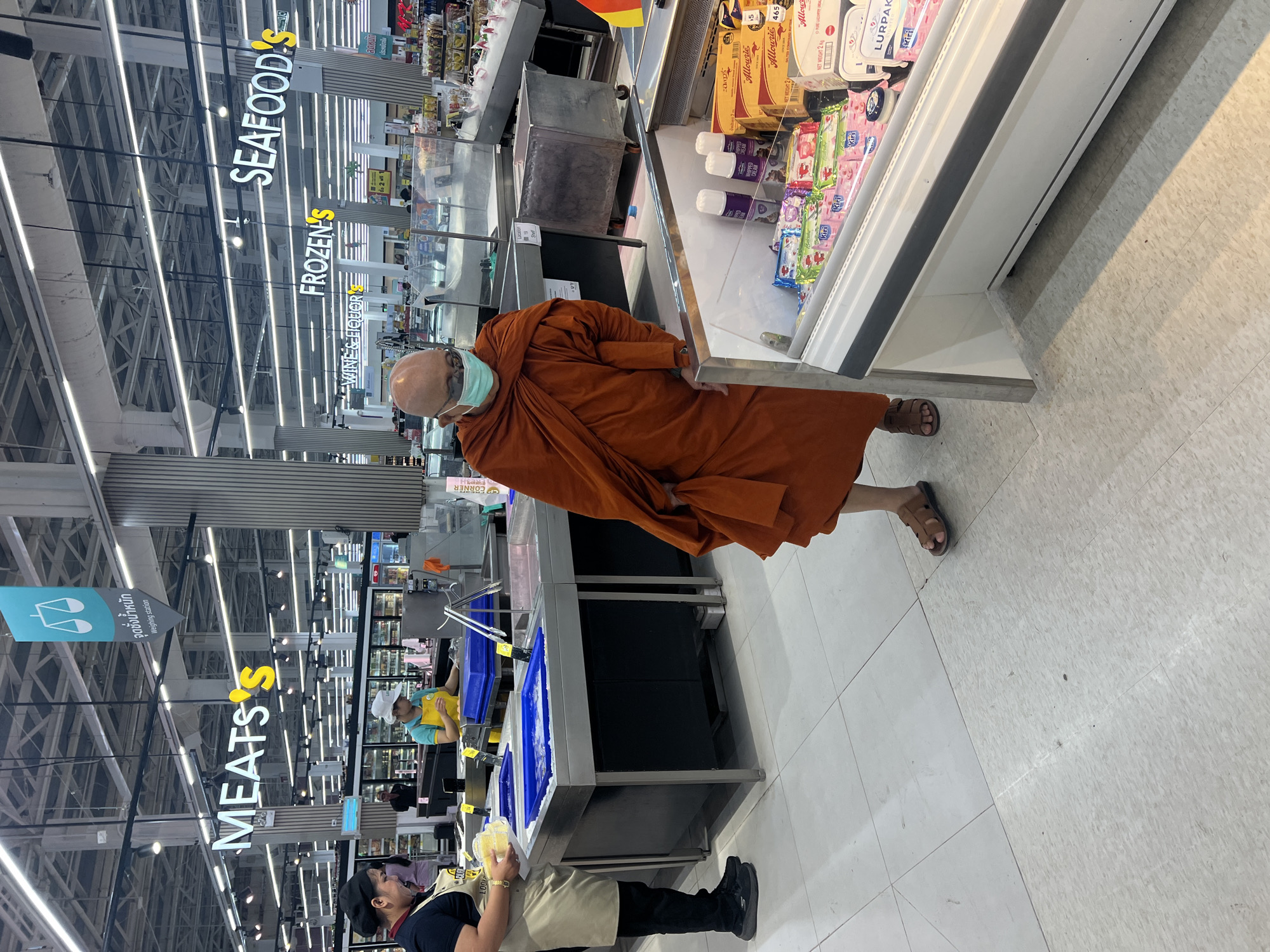 This image shows a person in a monk's robe and face mask standing next to a refrigerated display case in a supermarket. The person is wearing brown sandals. In the background, signs indicate the location of the "MEATS," "FROZEN'S," and "SEAFOOD'S" sections. Near the "MEATS" sign, a supermarket worker is visible. The refrigerated display case contains items such as butter and cheese. The image was taken in บ้านบางรักษ์, Thailand.