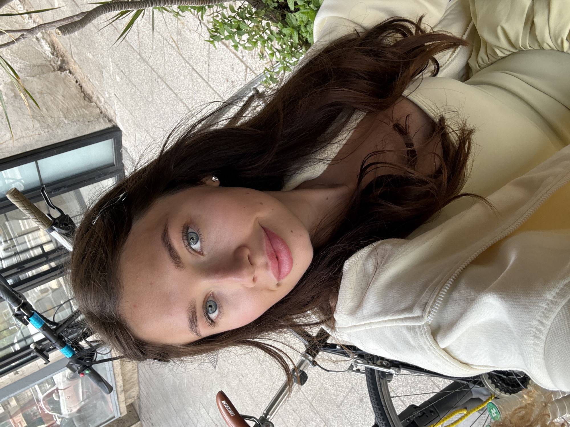 A young woman with long brown hair and blue eyes takes a selfie. She is wearing a light yellow jacket. A bicycle is partially visible in the foreground. The background includes a window and a potted plant. The location appears to be in an urban setting, specifically in Tel-Aviv, Israel. The presence of the bicycle and the young woman's casual attire suggests a possible activity of cycling.