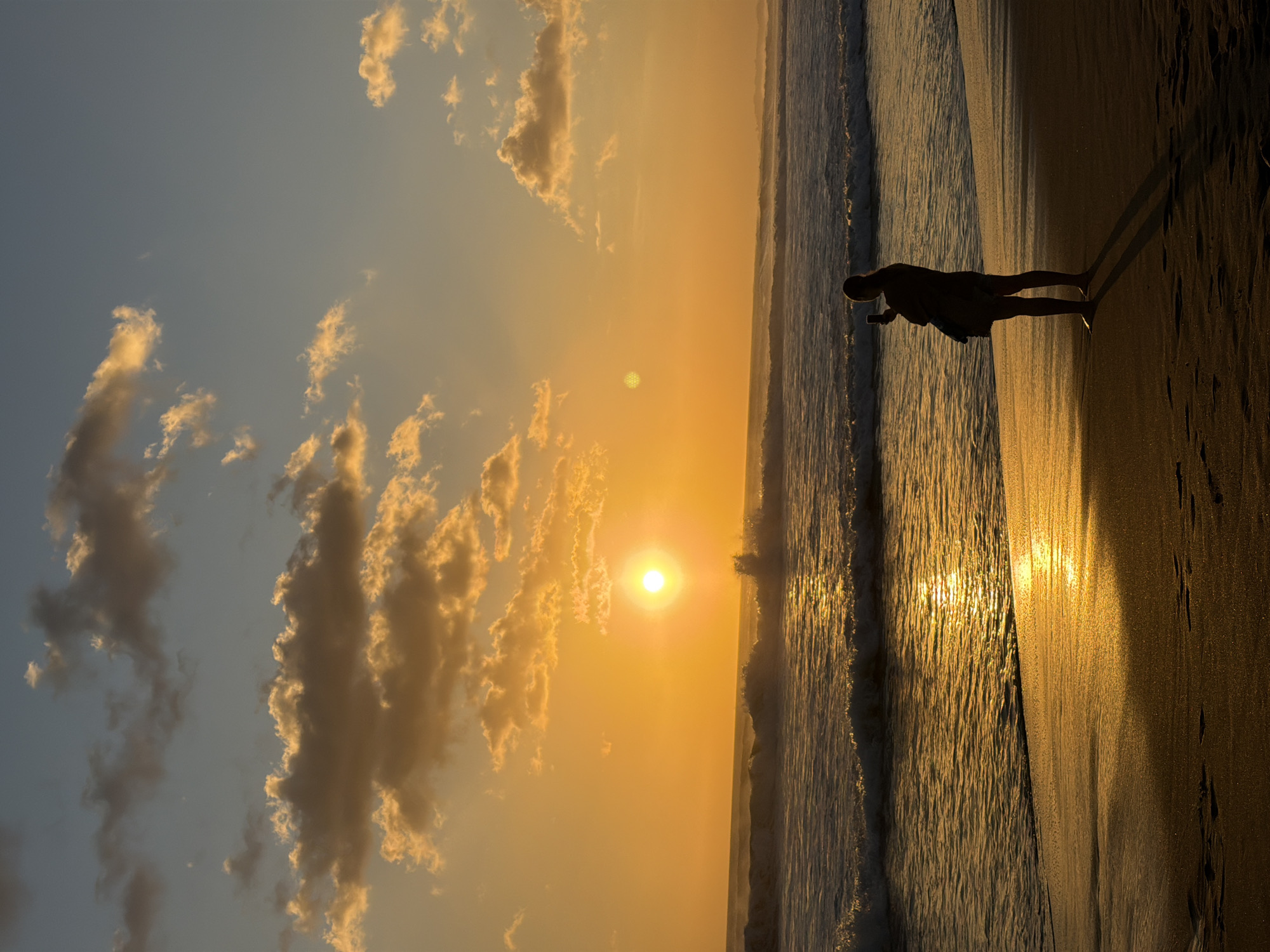A person stands on a sandy beach in North Kuta, Indonesia, silhouetted against a vibrant sunset. The sun, a bright orb, is low on the horizon, casting a golden glow across the sky and ocean. Clouds, tinged with orange and yellow, dot the sky. Waves crash gently onto the shore, their crests illuminated by the setting sun. The person, facing the ocean, appears to be taking a photograph with a mobile device. Footprints are visible in the wet sand near the water's edge. The overall atmosphere is serene and picturesque.
