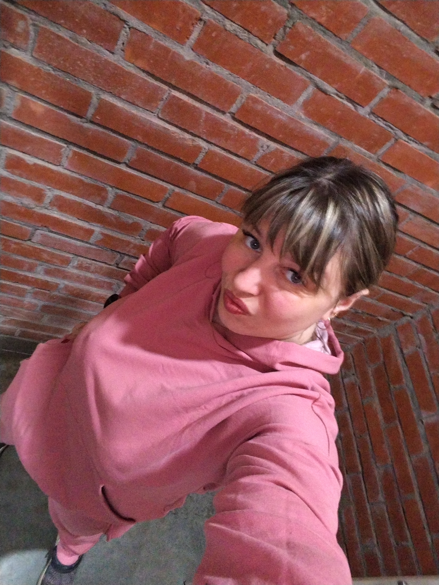 A high-angle selfie captures a woman in Rusanovo, Russia, presenting a playful expression with puckered, reddish-pink lips and wide blue eyes. She has short, styled hair with bangs and prominent blonde highlights. The woman is wearing a matching pink tracksuit, including a hooded sweatshirt with a large front pocket and pink pants. Dark sneakers are visible on her feet. A small, hoop-like earring is visible on her right ear. The background features a textured red brick wall with grey mortar lines, suggesting an indoor or sheltered outdoor location. A grey floor or ground surface is also visible in the lower part of the frame. The lighting is uniform, and there is no visible text in the image.