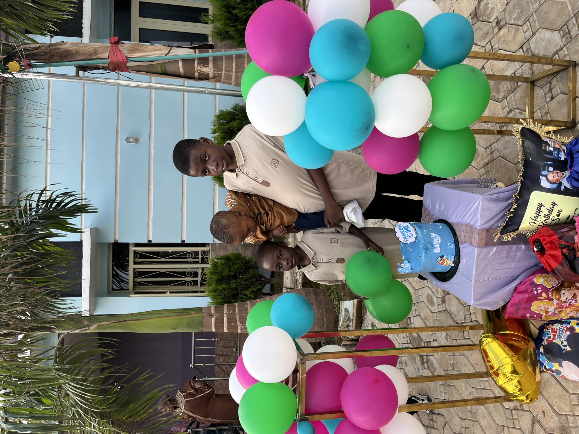 This image captures an outdoor birthday celebration in Jos, Nigeria. In the foreground, three males are present: an older boy, a younger boy, and an infant. The older boy, positioned on the right, wears a beige short-sleeved collared shirt with an 'N' logo on the left chest and dark trousers, holding an infant dressed in a brown long-sleeved top, blue jeans, and white sneakers. The infant faces away from the camera. To the left, a younger boy wears an identical beige collared shirt with an 'N' logo.
In the center-bottom, a light blue round birthday cake rests on a black base, placed on a small table draped in light purple fabric. The cake features "FAHEEM @ ONE" on a cloud-shaped topper and "Bluey" characters on its side. Around the table and on the light-colored interlocking paved ground, various gift bags, including Disney Princess and Mickey Mouse themes, are visible, along with a golden Mylar balloon. A black cushion with "Happy Birthday Son" and images is positioned on the bottom right.
Numerous balloons in green, white, magenta, and turquoise are arranged on two golden metallic vertical stands that flank the central area. Additional balloons are scattered on the ground. In the left background, an adult woman in a brown garment and head covering is partially visible, standing near a metal railing.
The background features a light blue two-story building with white horizontal trim lines and windows secured by white metal grates. Large palm trees with extensive green foliage frame the scene on both sides. A silver ladder, secured with red fabric, leans against the palm tree on the right. Smaller green bushes are visible near the building. The ground is uniformly covered with light-colored interlocking pavers.