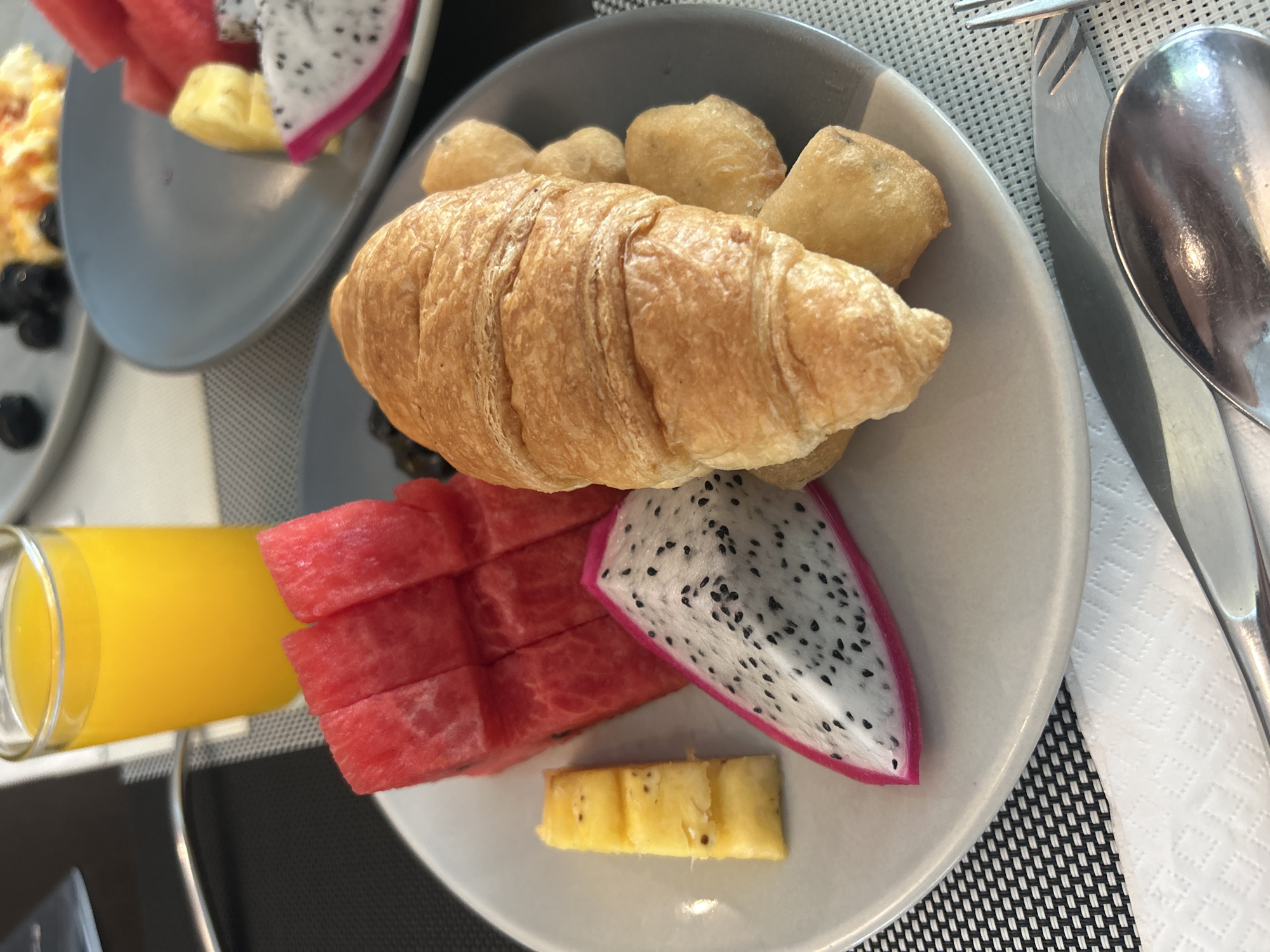 The image shows a breakfast setting. In the foreground, there is a gray plate containing a croissant, several fried dough balls, watermelon slices, dragon fruit slice, pineapple pieces, and an accompanying glass of orange juice. To the right of the plate are a fork and a spoon placed on a white napkin. Another plate is visible in the upper left corner with what appears to be scrambled eggs, blueberries, watermelon, dragon fruit, and pineapple. The background shows a textured surface, possibly a placemat, in a gray and white color scheme. While the image appears to be a breakfast scene, it does not provide any information to confirm the location is in Rawai, Thailand. Therefore, I cannot confirm the location is in the city of Rawai, Thailand.