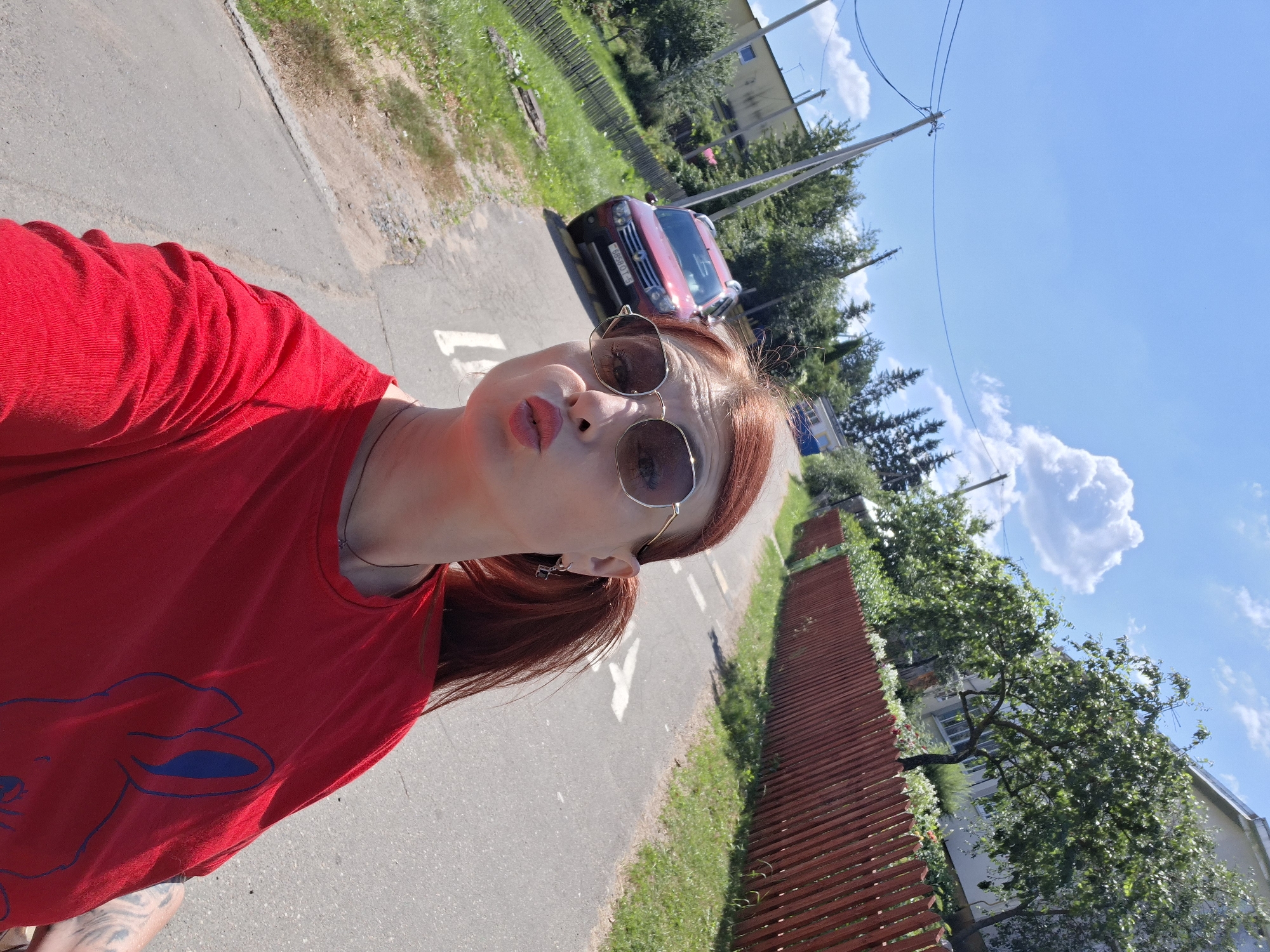 The image shows a woman with red hair, wearing a red t-shirt and sunglasses, taking a selfie outdoors on a sunny day. She is standing on a paved road with white markings. A red SUV is parked on the side of the road behind her. The background includes trees, a wooden fence, and a building. The location is identified as Забалоцкі сельскі Савет, Belarus.
