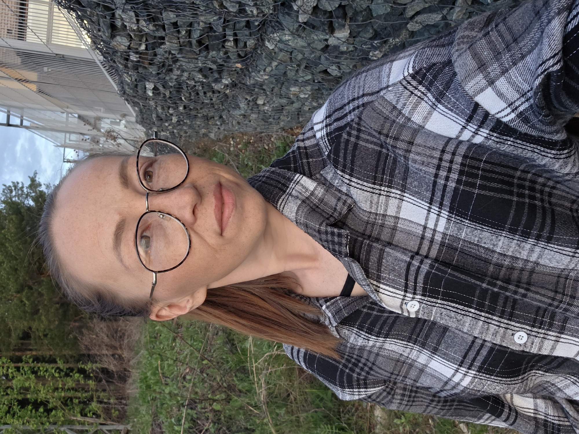 A woman with fair skin and brown hair, wearing round eyeglasses and a black and white plaid shirt with white buttons, is the main subject of the image. The location appears to be outdoors, with a gabion wall filled with grey rocks visible in the background, along with glimpses of greenery suggesting trees and vegetation. A white fence is visible in the distance. Based on visual data, it is impossible to conclusively determine the exact location in Chaikovsky, Russia, without more information.