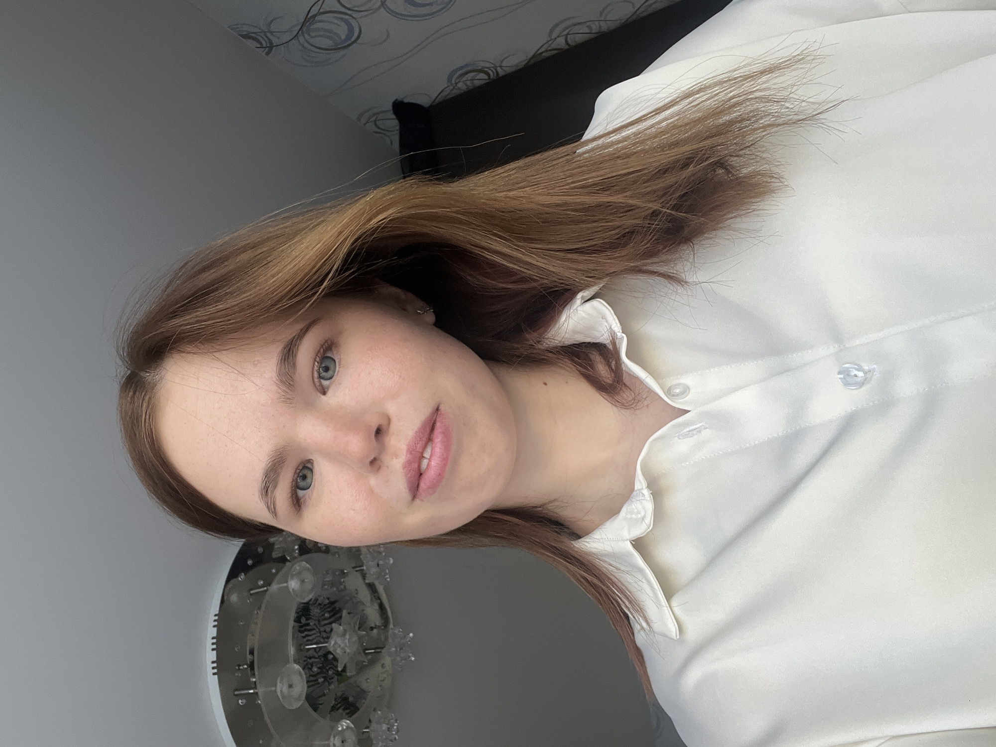 This image captures a young female subject positioned indoors, within a private residence in Chaikovsky, Russia. The subject, in her late teens or early twenties, has fair skin with visible freckles on her nose and cheeks, light blue-grey eyes, and light brown hair parted centrally. She is wearing a white collared button-down shirt with the top two buttons unfastened. A small silver hoop earring is visible in her left ear. Her lips are slightly parted, revealing her upper teeth. The background features a light grey ceiling from which a portion of a multi-tiered chandelier with clear elements is visible in the upper left. The wall on the right side of the frame is covered with patterned wallpaper displaying grey and white swirling designs, while the wall on the left is a solid light grey. A dark, undefined object is partially visible behind the subject's right shoulder against the patterned wallpaper.