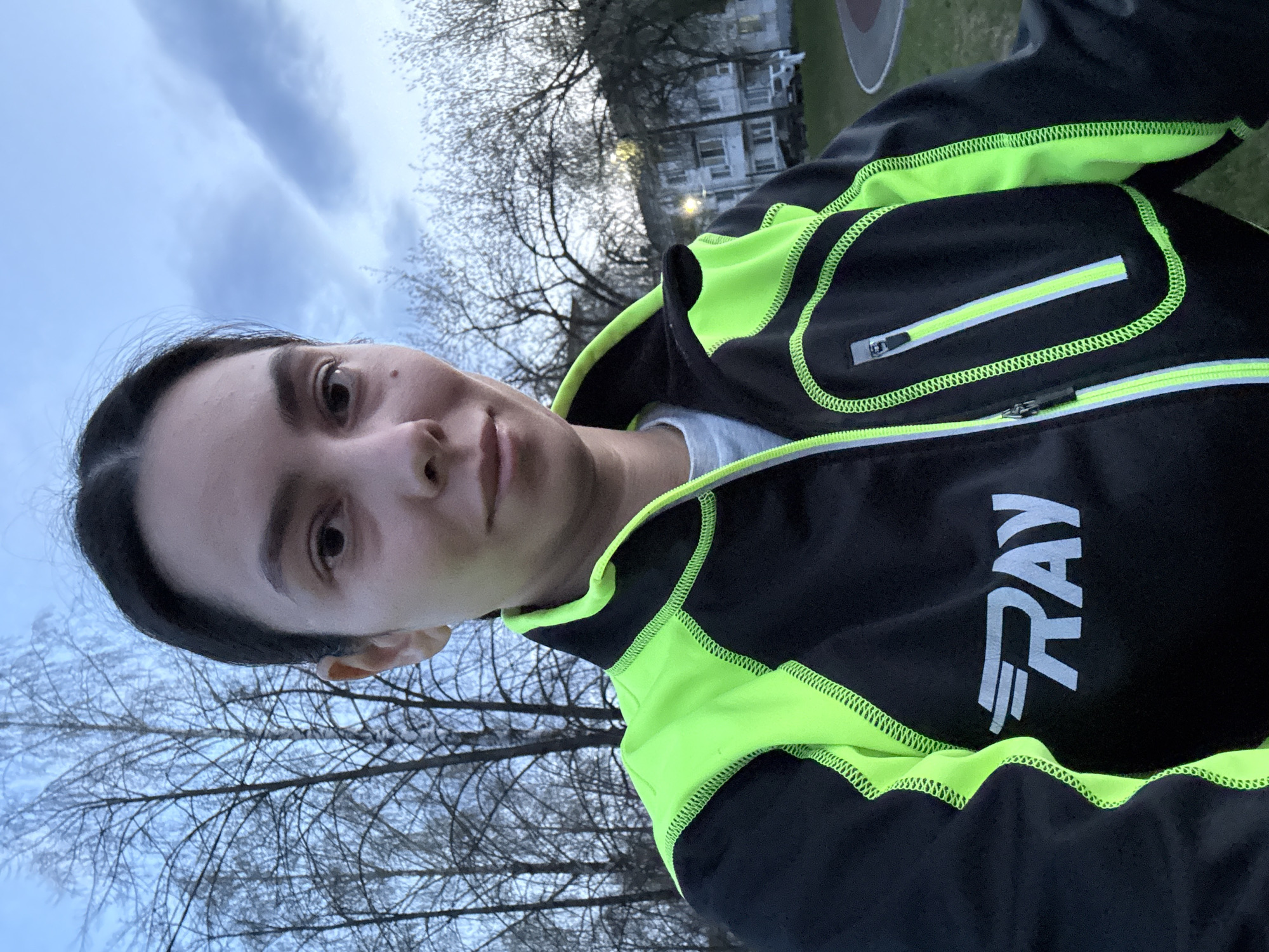 A selfie taken by a woman wearing a black and neon yellow jacket with the word "RAY" printed on it. The background shows bare trees, a building with windows, and a cloudy sky. Based on the presence of the building and park-like setting, the photo appears to be taken in an urban environment, possibly in the city of Chaikovsky, Russia.