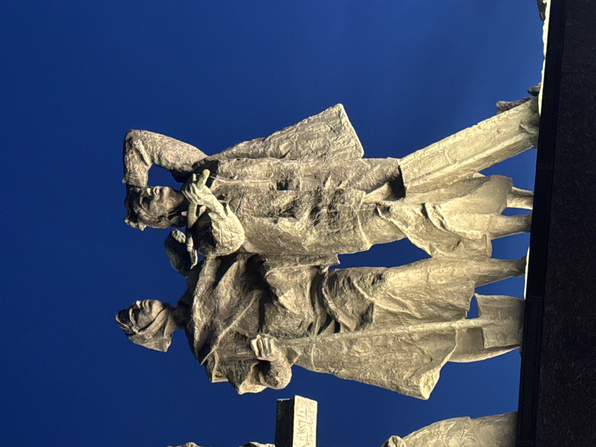 The image captures a detailed, low-angle perspective of a section of the Monument to the Heroic Defenders of Leningrad, located at Victory Square (Ploshchad Pobedy) in Saint Petersburg, Russia. The scene is illuminated by artificial light against a dark, clear blue sky, suggesting a nighttime or twilight setting.
The primary subjects are two monumental bronze figures standing side-by-side on a dark, stone plinth, visible at the bottom of the frame. The statues exhibit a textured, rugged surface.
On the left, a female figure is depicted, wearing a headscarf or hood and a long, flowing garment. She holds a vertical implement, possibly a shovel handle or staff, in her left hand, which extends downwards. Her gaze is directed upwards and slightly to the right. A shovel blade is distinctly visible near her lower legs. To her immediate left, a rectangular object, possibly part of another monument element, displays partially visible numerals that appear to be "1974".
To the right of the female figure, a male figure is presented in a vigilant stance. He wears a hat or head covering and a heavy coat. His right arm is raised, with his hand positioned above his brow, as if shielding his eyes to scan the distant horizon or sky. His left hand holds a long, slender object, consistent with a rifle or a similar tool, which rests vertically against his body. His head is also turned upwards and to the right, mirroring the female figure's direction of gaze.
The artificial lighting casts a warm, golden-brown hue on the statues, highlighting their sculptural details and creating strong contrasts against the deep blue background. No other elements or individuals are distinctly visible beyond the monument and the sky.