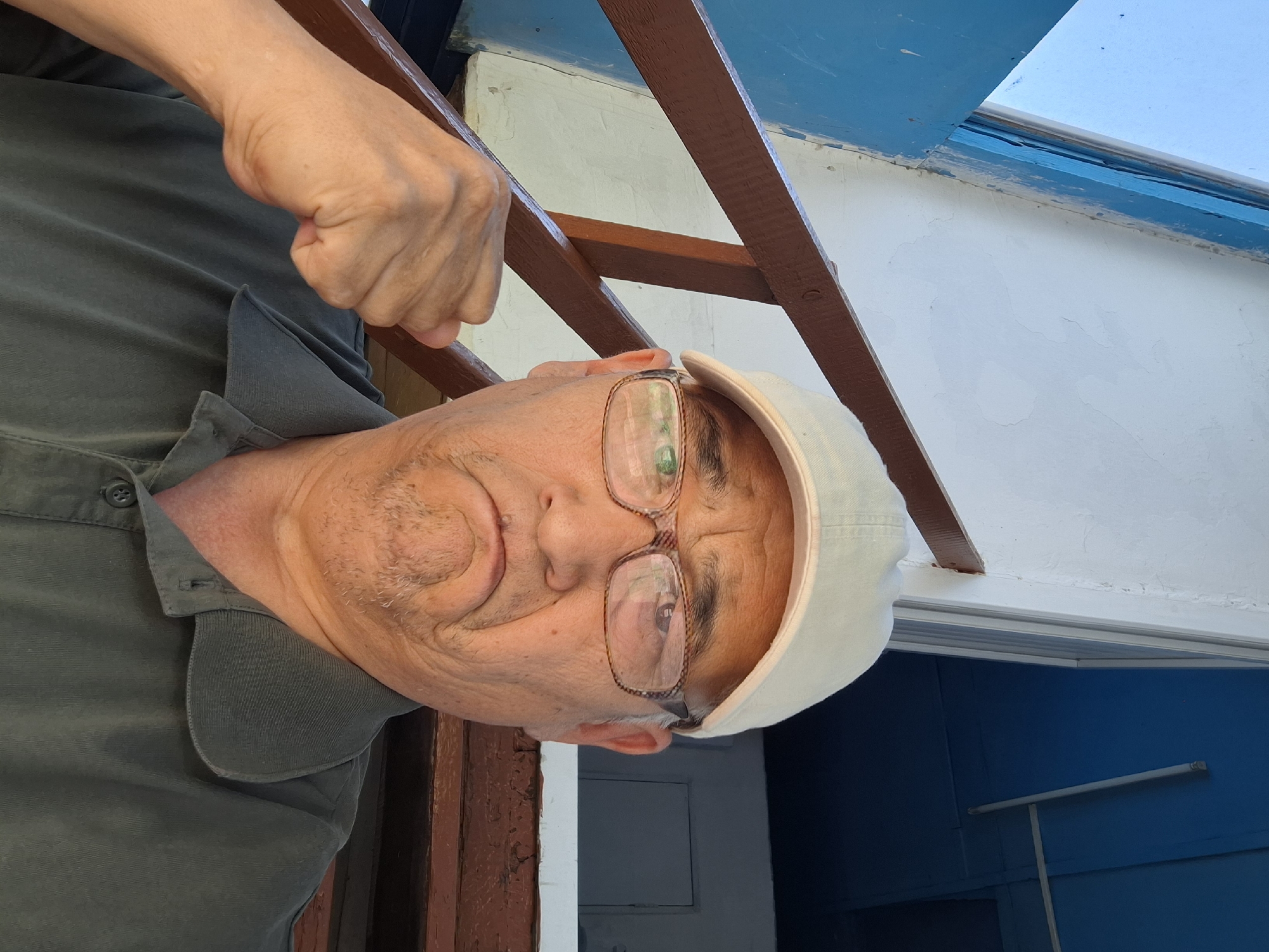 A man is pictured in an outdoor setting, possibly in the city of Almaty, Kazakhstan. He is wearing a light-colored baseball cap and glasses. His expression is one of displeasure, with his mouth turned down and his brow furrowed. His right fist is raised, clenched in front of his chest. He is wearing a dark green polo shirt. Behind him, a wooden railing and a white wall are visible. The background also suggests an urban environment with hints of blue structures. The lighting indicates it is daytime.