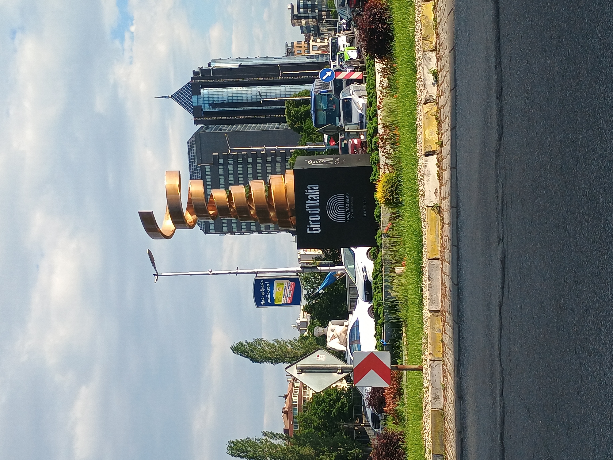 The image shows a street scene with a large golden sculpture of a spiral shape located next to a sign that says "Giro d'Italia" in the city of Plovdiv, Bulgaria. In the background are several modern high-rise buildings, and a line of parked vehicles. A paved road runs along the right side of the image, adjacent to a curb with grass and shrubbery. Road signs and street furniture are also visible, along with trees and other greenery. The sky is partly cloudy.