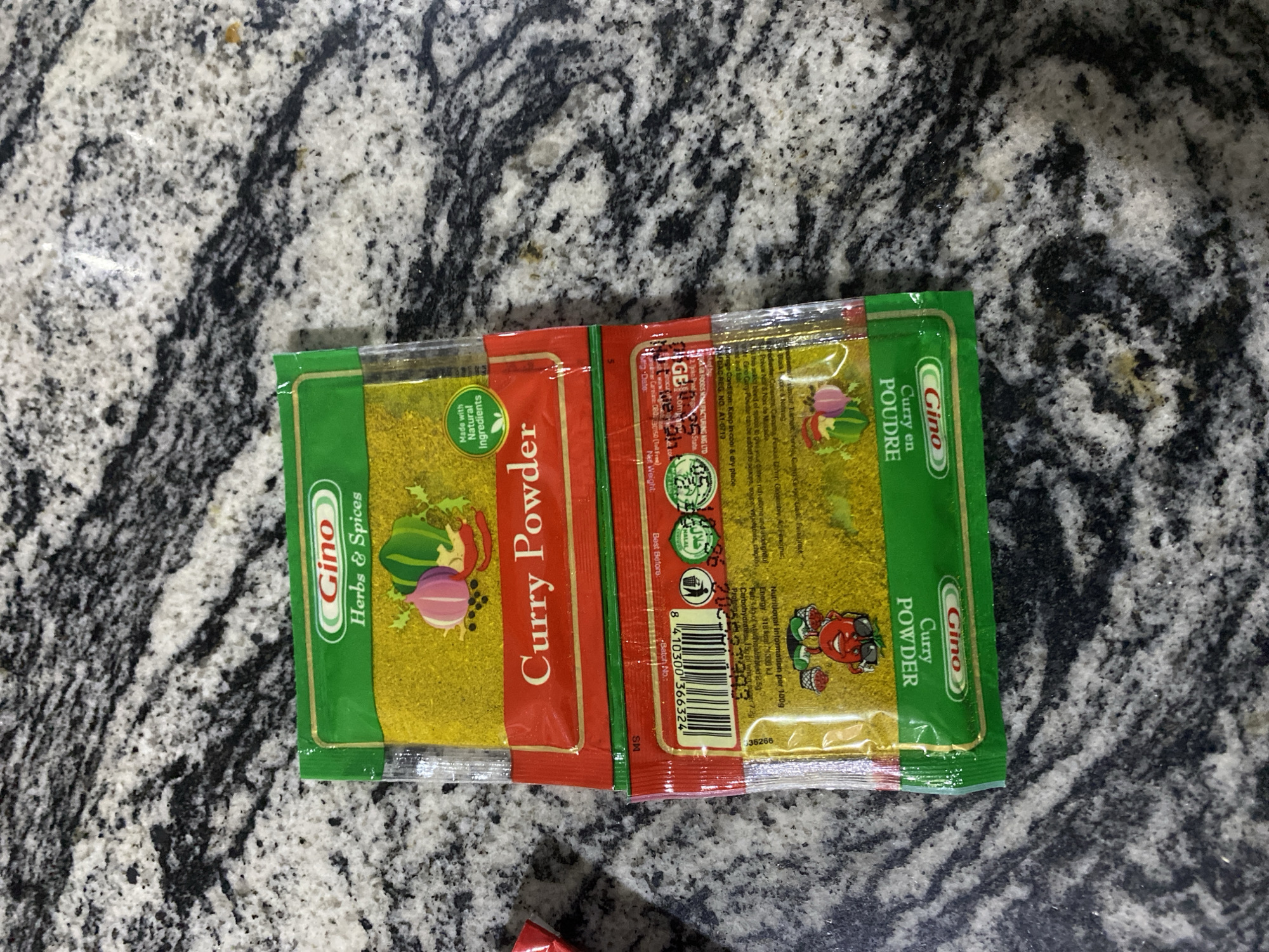 A close-up, overhead shot shows two sealed packets of Gino Curry Powder stacked on a granite countertop. The top packet is bright green with "Gino Herbs & Spices" printed in white and red lettering. Below that, a red band displays "Curry Powder" in white text. A graphic of a red chili pepper, a purple onion, and green peppers sits above the text. The bottom packet, also green, shows "Gino Curry Powder" in white lettering and "Curry en Poudre" in smaller white text. Both packets are partially filled with a yellow powder. The background is a mottled grey and black granite surface. The scene is static and there are no people or animals visible. The location is not discernible from the image.