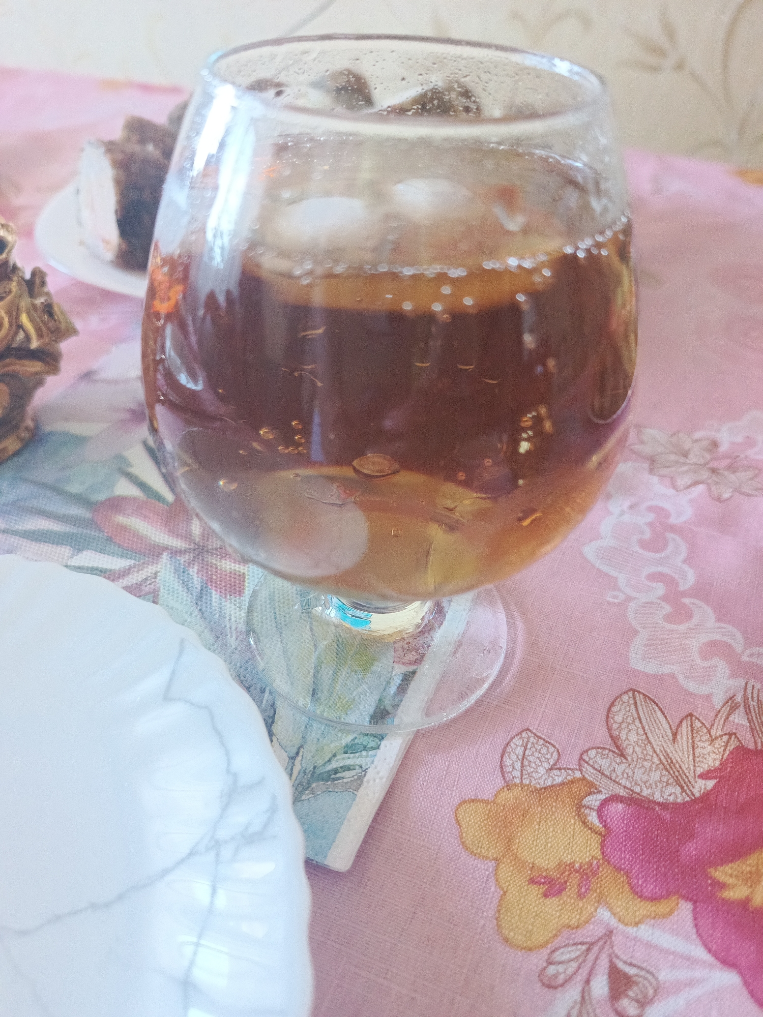 A close-up, overhead shot shows a glass of amber liquid with ice cubes and condensation on the glass, sitting on a pink tablecloth with a floral pattern. To the left, a white plate with what appears to be a piece of cake is visible. The background is blurry, with hints of a wall and other objects. The lighting is soft and natural. The scene appears to be set indoors, possibly in a dining area or kitchen. The image is a still life, focusing on the beverage and accompanying food item.