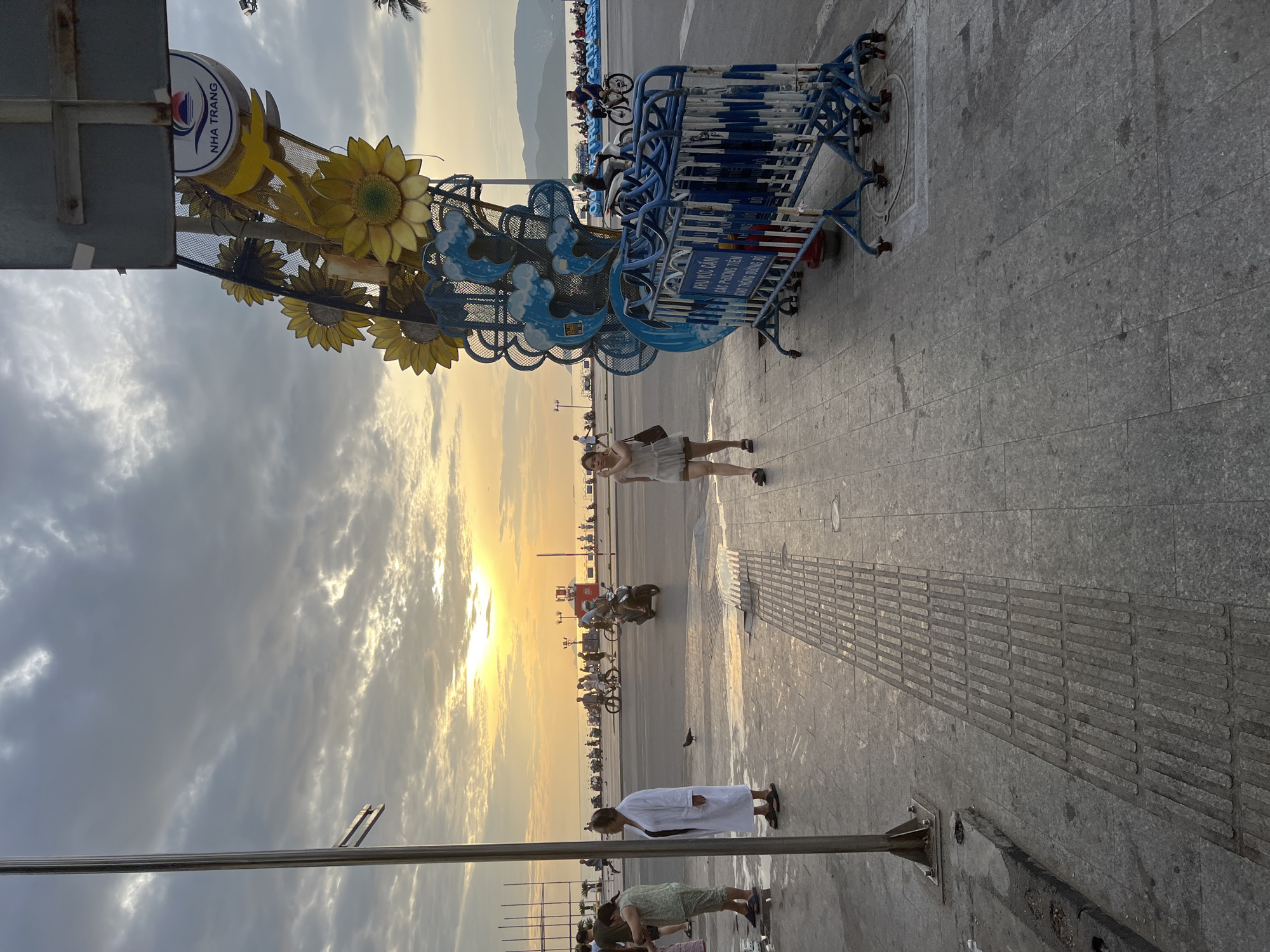 The image captures a street scene in Nha Trang, Vietnam, during sunset. The sky is filled with dramatic clouds, tinged with the warm hues of the setting sun. In the foreground, a woman in a light-colored dress walks on a paved sidewalk. To her right, a temporary blue and white barricade stands, with a decorative structure featuring sunflowers and wavy patterns behind it. A sign with Vietnamese text is visible on the barricade. Further down the street, several people are walking and riding motorcycles and bicycles. In the background, a body of water, likely the sea, is visible, with hills on the horizon. The overall atmosphere is one of a bustling coastal city at dusk.