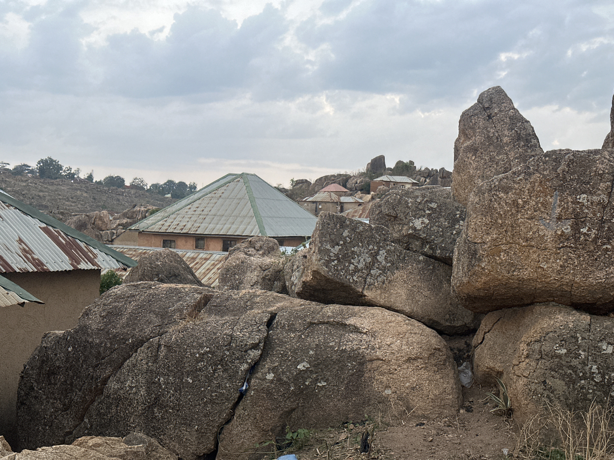The image shows a scene in Jos, Nigeria, characterized by large granite boulders dominating the foreground. A cluster of structures with corrugated metal roofs is situated behind and between the rocks. One building features a distinct pyramid-shaped roof. The sky is overcast with grey clouds. Dry vegetation is visible at the base of the rocks.