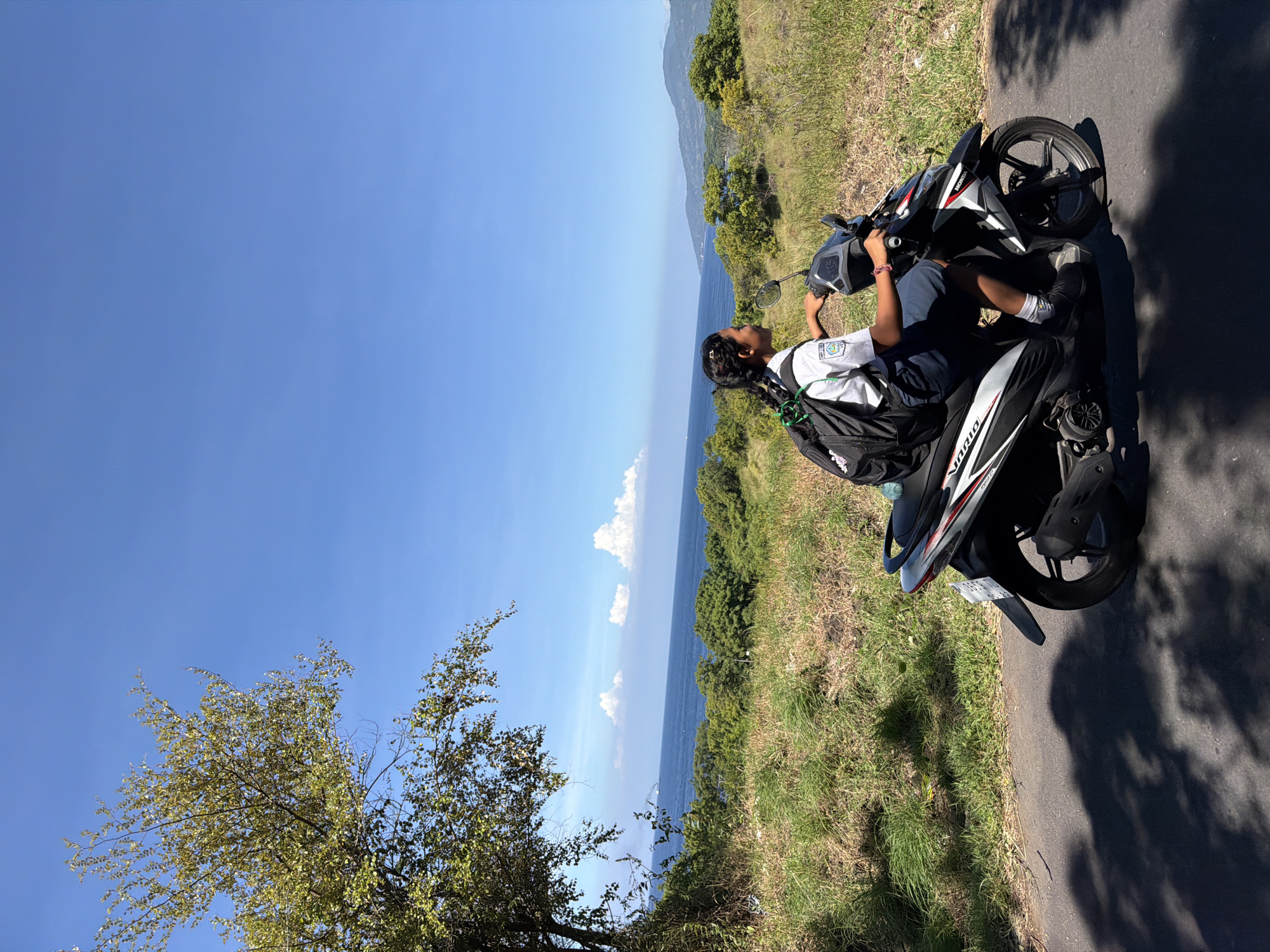 A person sits on a scooter parked on the side of a road in Kubu, Indonesia. The person has dark hair and skin and is wearing a white shirt with a logo on the left breast, dark pants, and white socks with dark sneakers. They have a black backpack on their back. The scooter is a white and black Honda Vario. The road is asphalt and has a shadow cast upon it. To the left of the road is green grass and foliage, with a tree in the foreground. In the distance, the ocean is visible with a mountainous coastline. The sky is blue with white clouds.