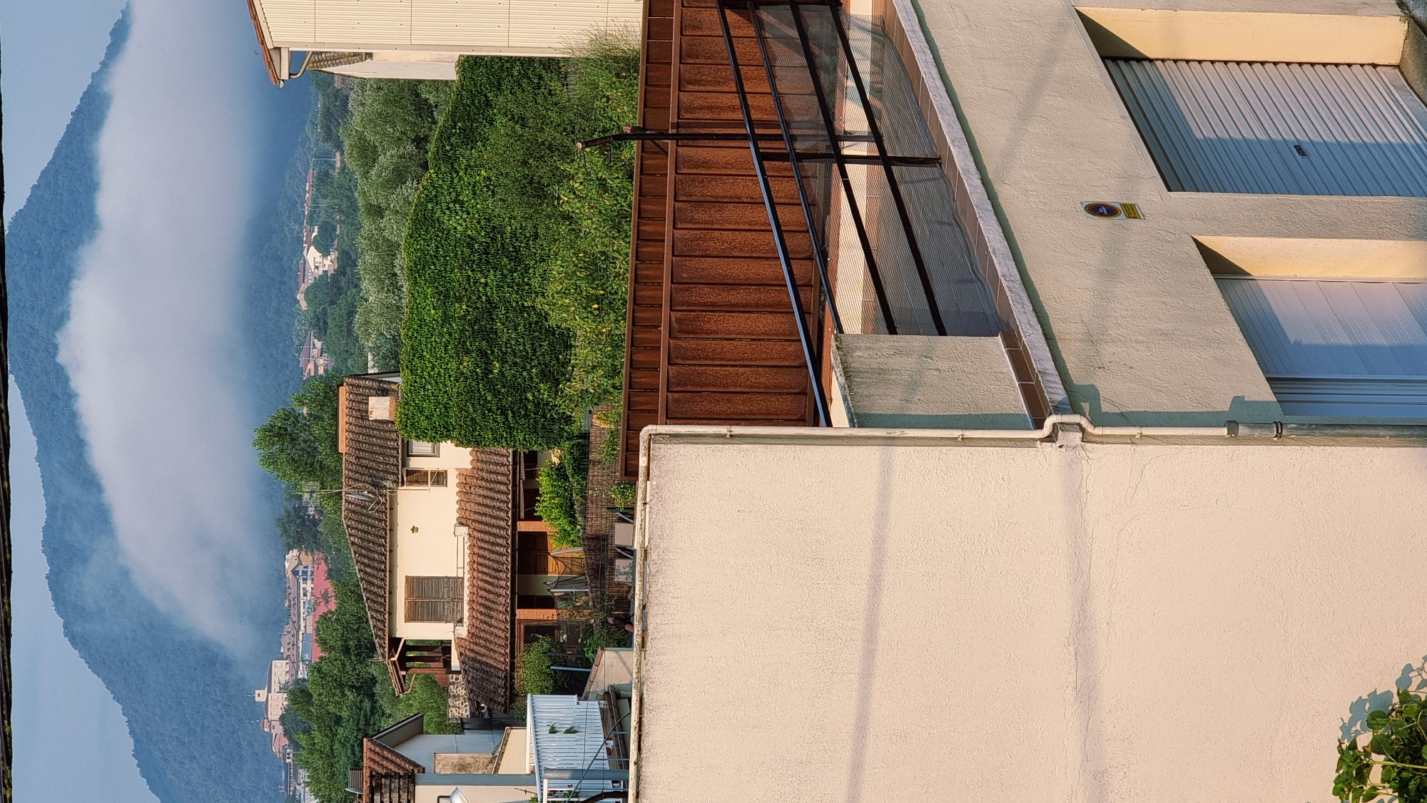 The image depicts a residential area in Sant Joan Les Fonts, Spain, viewed from an elevated perspective. In the foreground, a light-colored, textured wall of a building dominates the lower left quadrant. Adjacent to this is another building with a cream-colored facade, featuring two garage doors with rolled-up shutters and a prominent vertical white pipe. A yellow circular sign with a symbol is affixed to this building. The middle ground reveals a rusty corrugated metal fence topped with a black metal railing and mesh, separating different property levels. Behind this fence, a dense green hedge forms a natural barrier, concealing further structures. Further back, traditional Spanish-style houses with terracotta tiled roofs are visible, surrounded by lush green trees. In the distance, the silhouette of a mountain range is shrouded in a thick layer of mist, with some buildings peeking through at its base. The lighting suggests it is either early morning or late afternoon, casting long shadows.