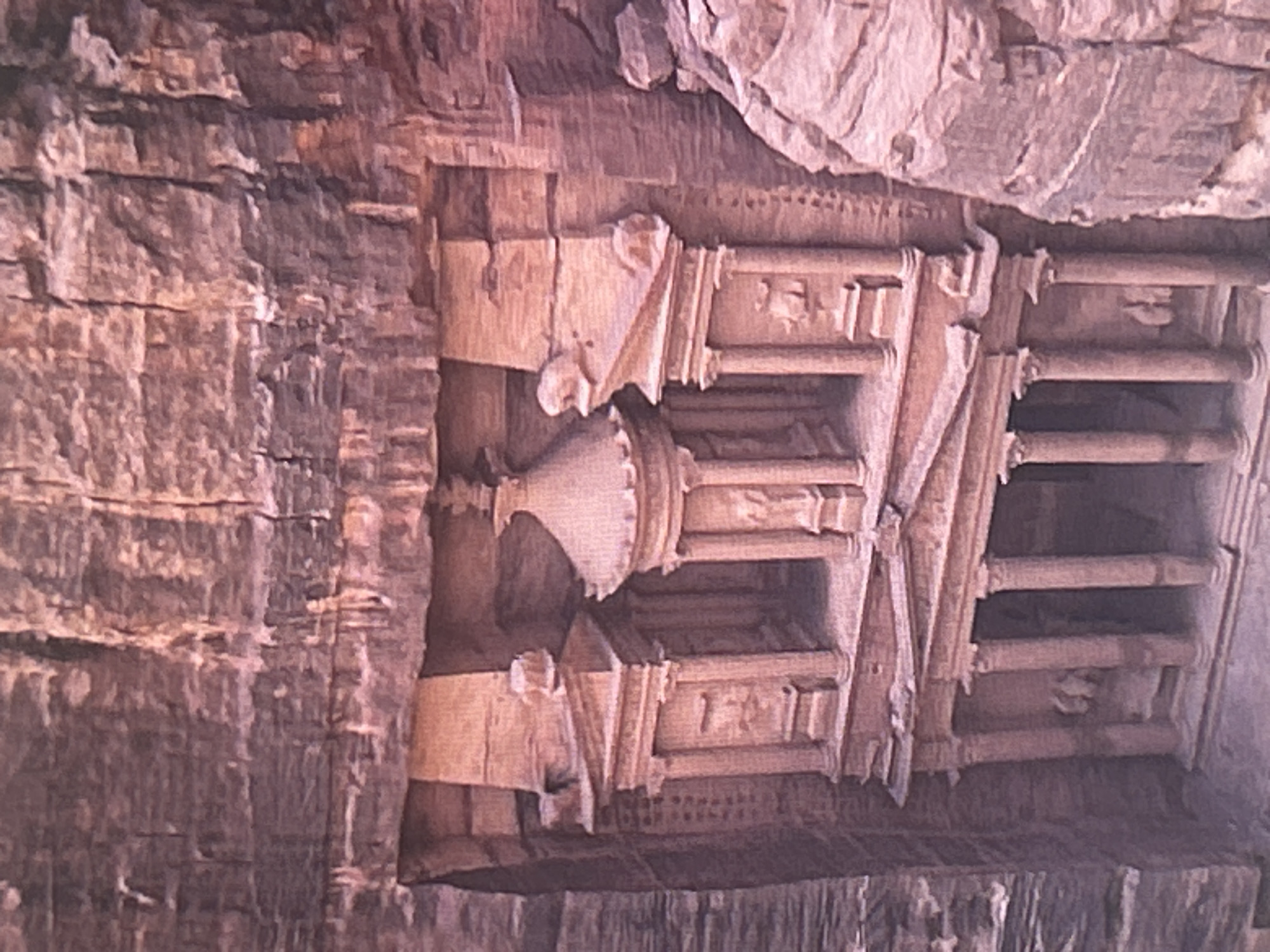 The image shows an aerial view of a large, ancient structure carved into a sandstone cliff face. The structure has a grand, ornate facade with columns, pediments, and niches. The rock itself is a reddish-brown color and appears weathered, with visible striations and cracks. The overall impression is one of historical significance and monumental architecture. This location is Petra, Jordan, not Sofia, Bulgaria.