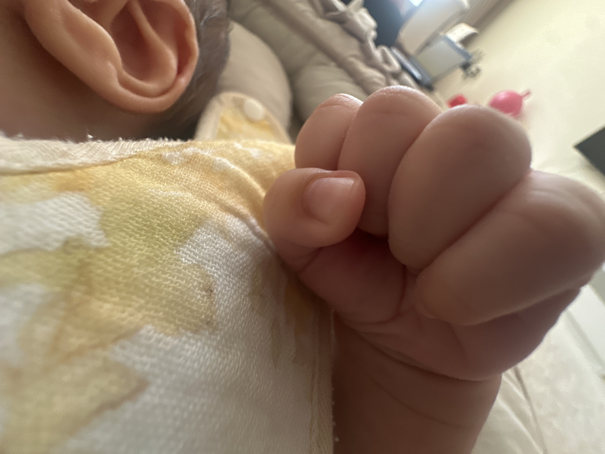 A close-up, low-angle shot shows the tightly clenched fist of a baby, resting on a light-colored cloth with a yellow floral pattern. To the left of the fist, a baby's ear is partially visible, showing the curves and folds of its outer structure. The background is blurred, with hints of furniture and a pink object, possibly a toy, in the distance. The scene is indoors, with soft lighting. There is no information to determine the location in the city of Masazir, Azerbaijan.