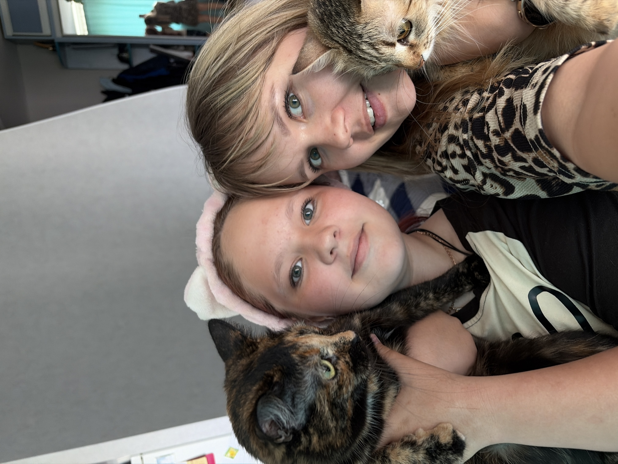 Two light-skinned females are posing with cats. The woman on the top is wearing a cheetah-print shirt. She is holding a tabby cat. The girl on the bottom is wearing a pink headband with ears. She is holding a calico cat. The location appears to be indoors. No specific details indicate the scene's location to be Chaikovsky, Russia.