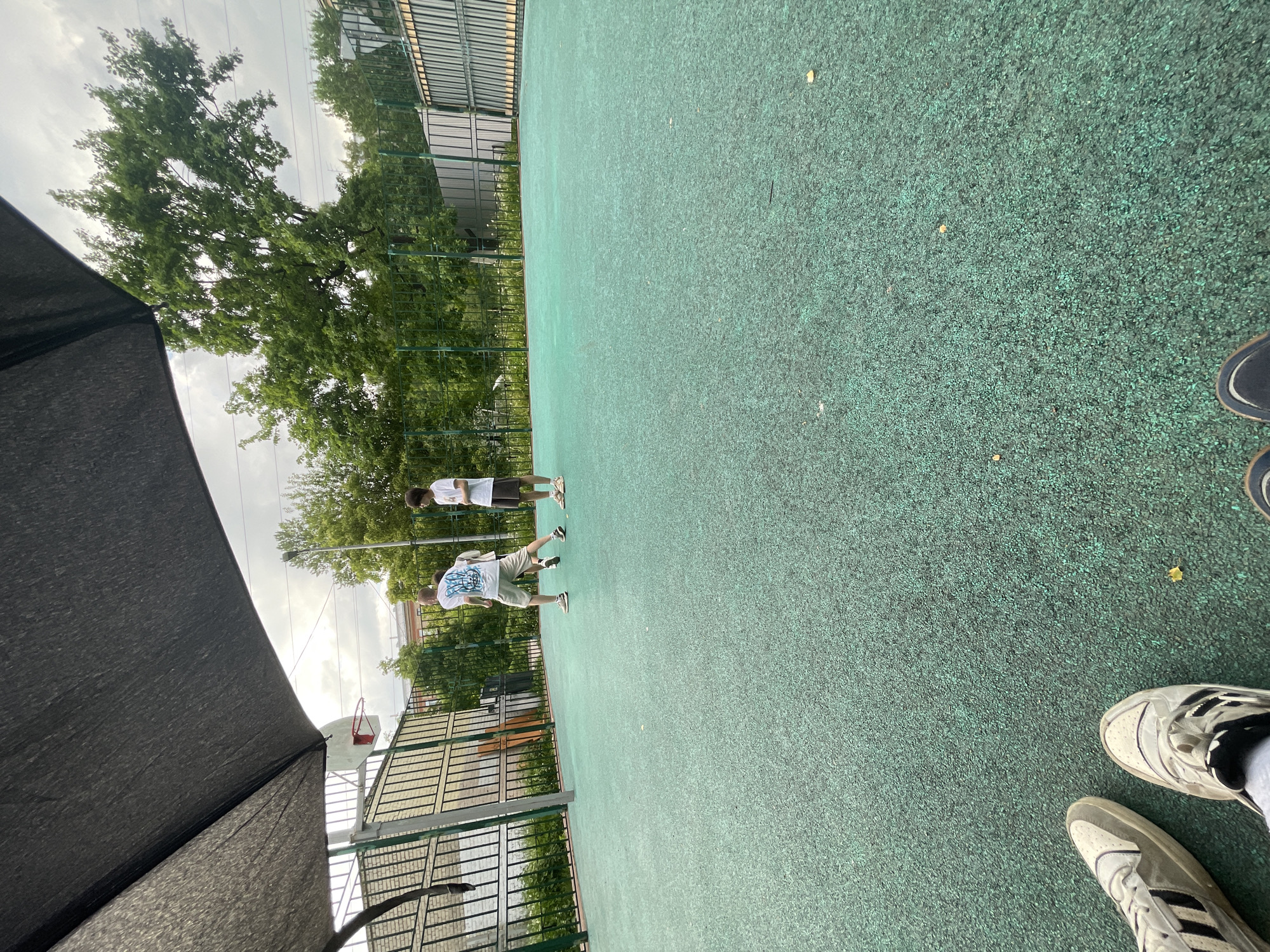 This image depicts an outdoor sports court on an overcast day. The main subject is a textured, green court surface, likely made of rubberized material or asphalt. Two young men are standing on the court, casually dressed in t-shirts and shorts. One, positioned closer to the viewer on the left, wears a white t-shirt with a blue graphic or text (which is unreadable) and light-colored shorts. The other, further back and to the right, wears a plain white t-shirt and darker shorts.
In the bottom right corner, the feet and lower legs of a third person, presumably the photographer, are visible, wearing white sneakers with black stripes and dark blue or black shoes.
The court is enclosed by a tall metal fence, beyond which dense green trees and parts of buildings can be seen against a cloudy sky. A red basketball hoop and backboard are partially visible on the far left, behind a large, dark fabric structure that extends into the foreground, possibly a sunshade or tent. The setting suggests a casual, outdoor activity, possibly basketball or a general gathering, occurring during the daytime under cloudy conditions. The overall atmosphere appears relaxed. The context provided indicates the location is Saint Petersburg, Russia.