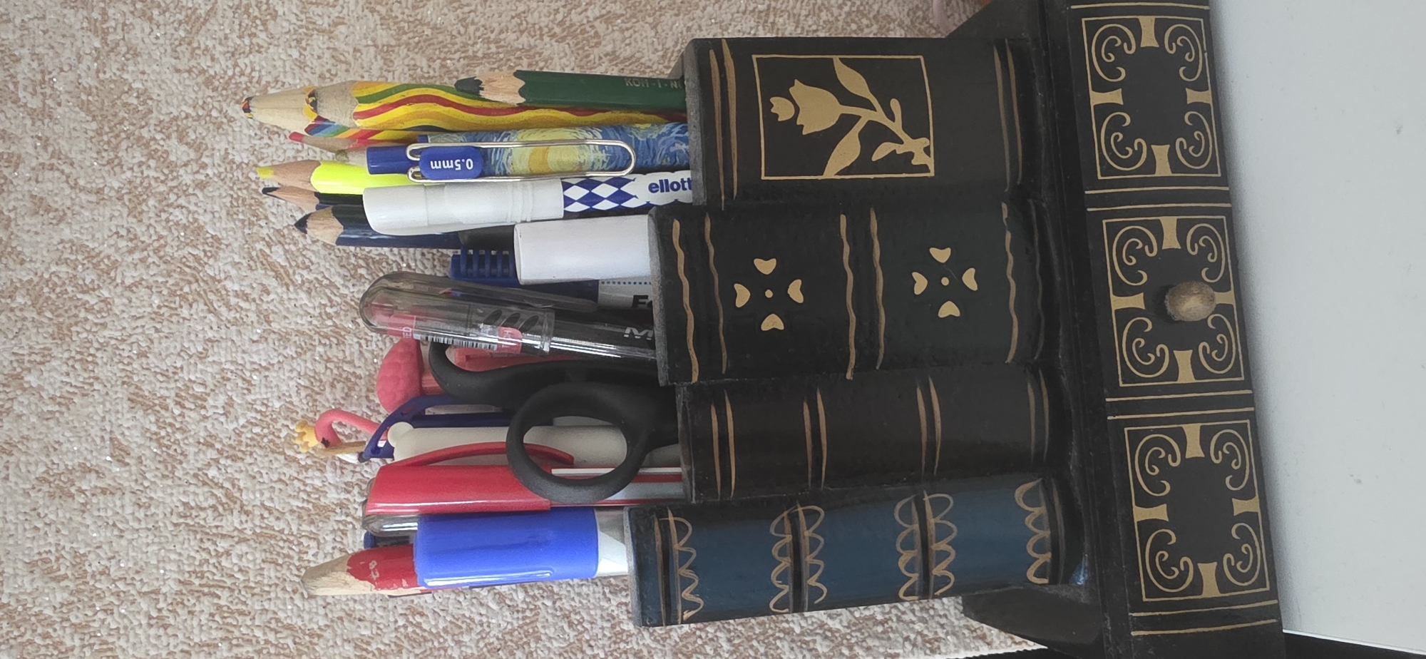 The image shows a desk organizer shaped like stacked books, filled with pens and pencils. The organizer is dark, with decorative gold trim and what appears to be a small drawer with a wooden knob. The items within the organizer include colored pencils, pens, and what looks like a small plastic flamingo toy. The background is a textured, light-colored wall. There are no people or animals present in the image. The scene is an indoor setting, likely a desk or shelf. There is no information or visual evidence in the image that can be used to determine its location in the city of Sumgayit, Azerbaijan.